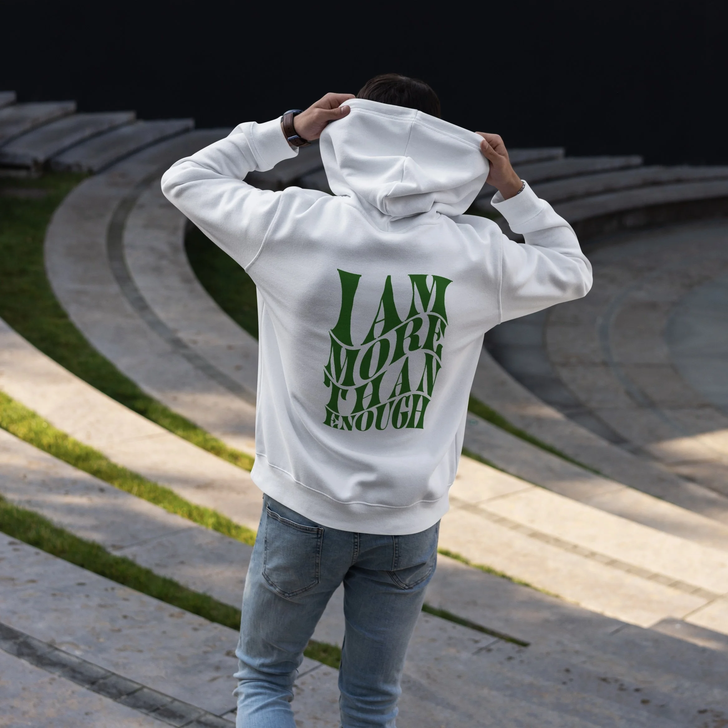 Person in a white hoodie and jeans adjusting their hood while standing outdoors against a dark sky, with curved concrete benches and grass in the background.
