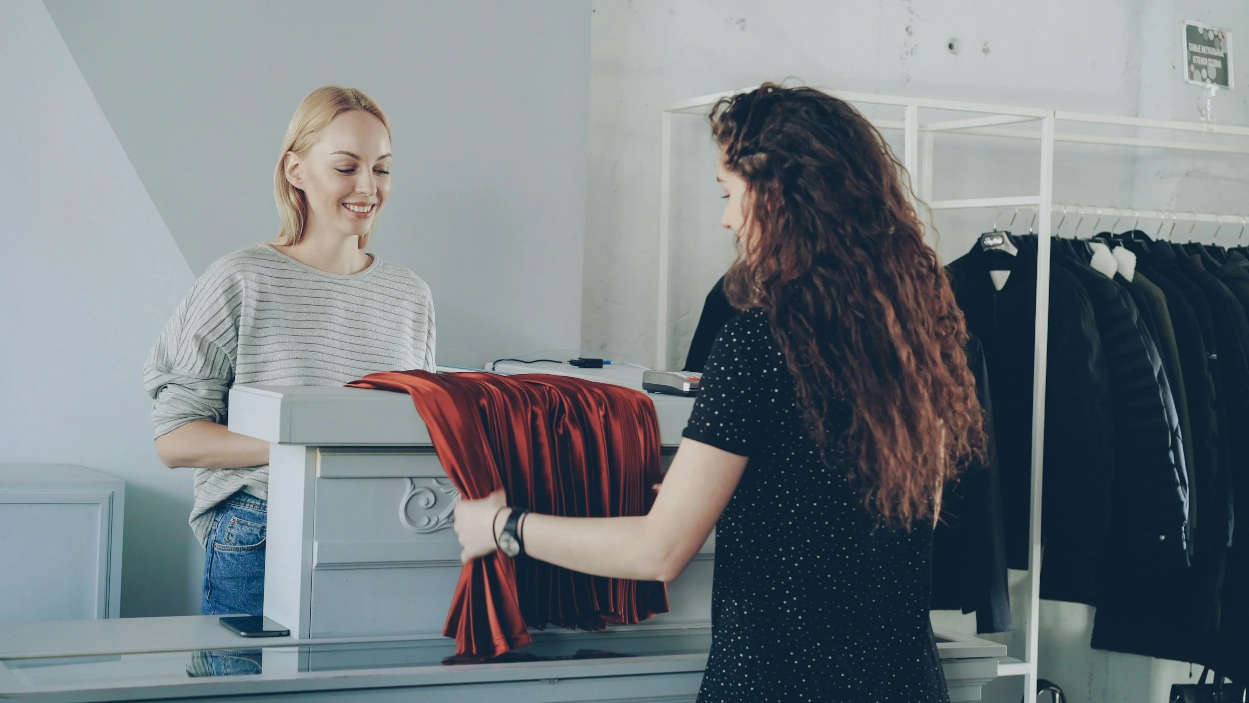 A woman with long, curly red hair wearing a black dress with white polka dots is receiving a red, silky dress as a gift from a blonde woman wearing a light gray striped sweater at a clothing store checkout counter.