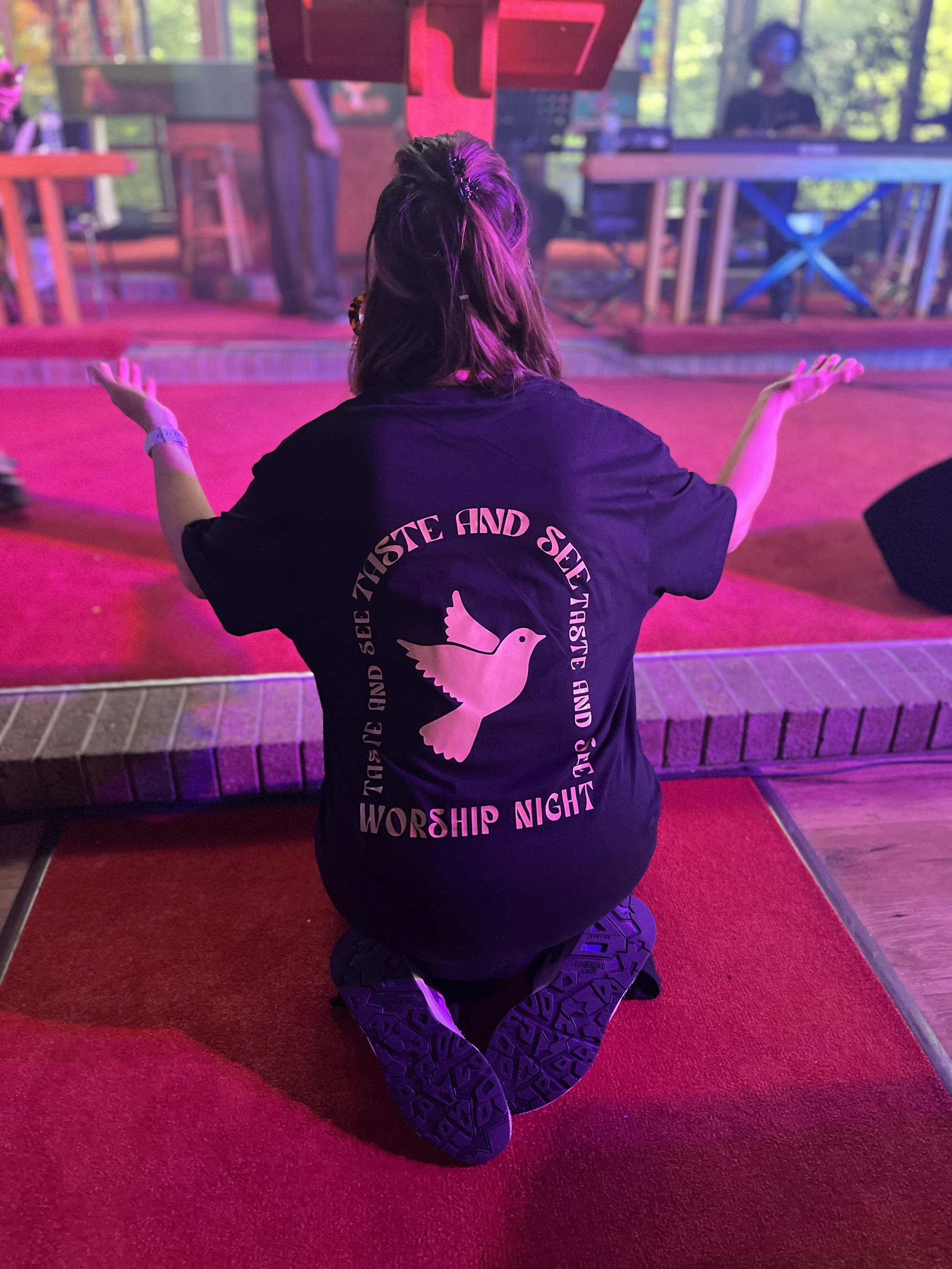 A person kneeling on a red carpet in a church or worship space, facing away, with arms raised. They are wearing a black t-shirt with a dove and circular text that reads "Worship Night" on the back. The space has a stage with people, musical equipment