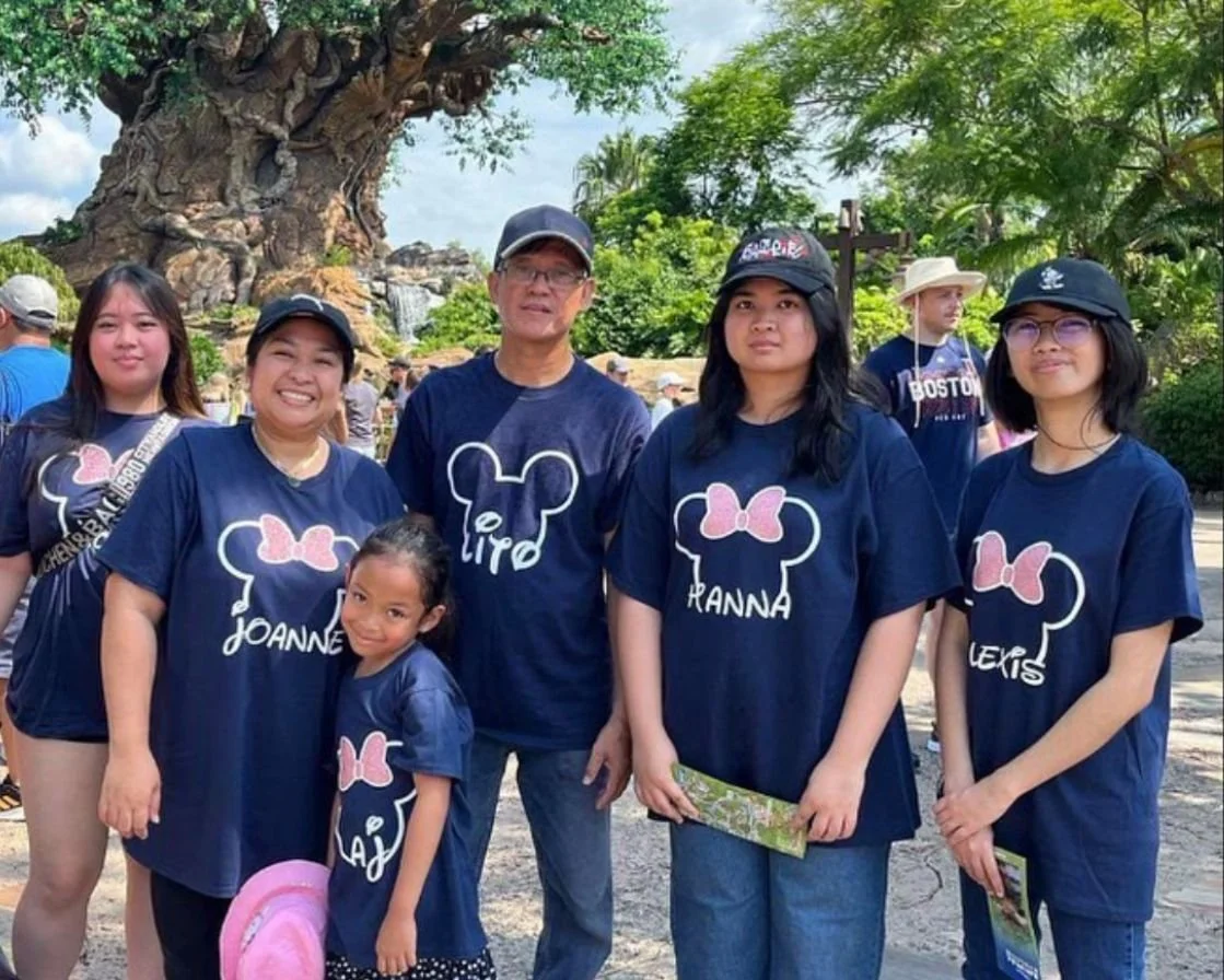 A family of six standing outdoors, all wearing matching navy blue shirts with a pink bow and bear face outline, with personalized names. They are near a large tree and surrounded by other people, with green trees and a blue sky in the background.