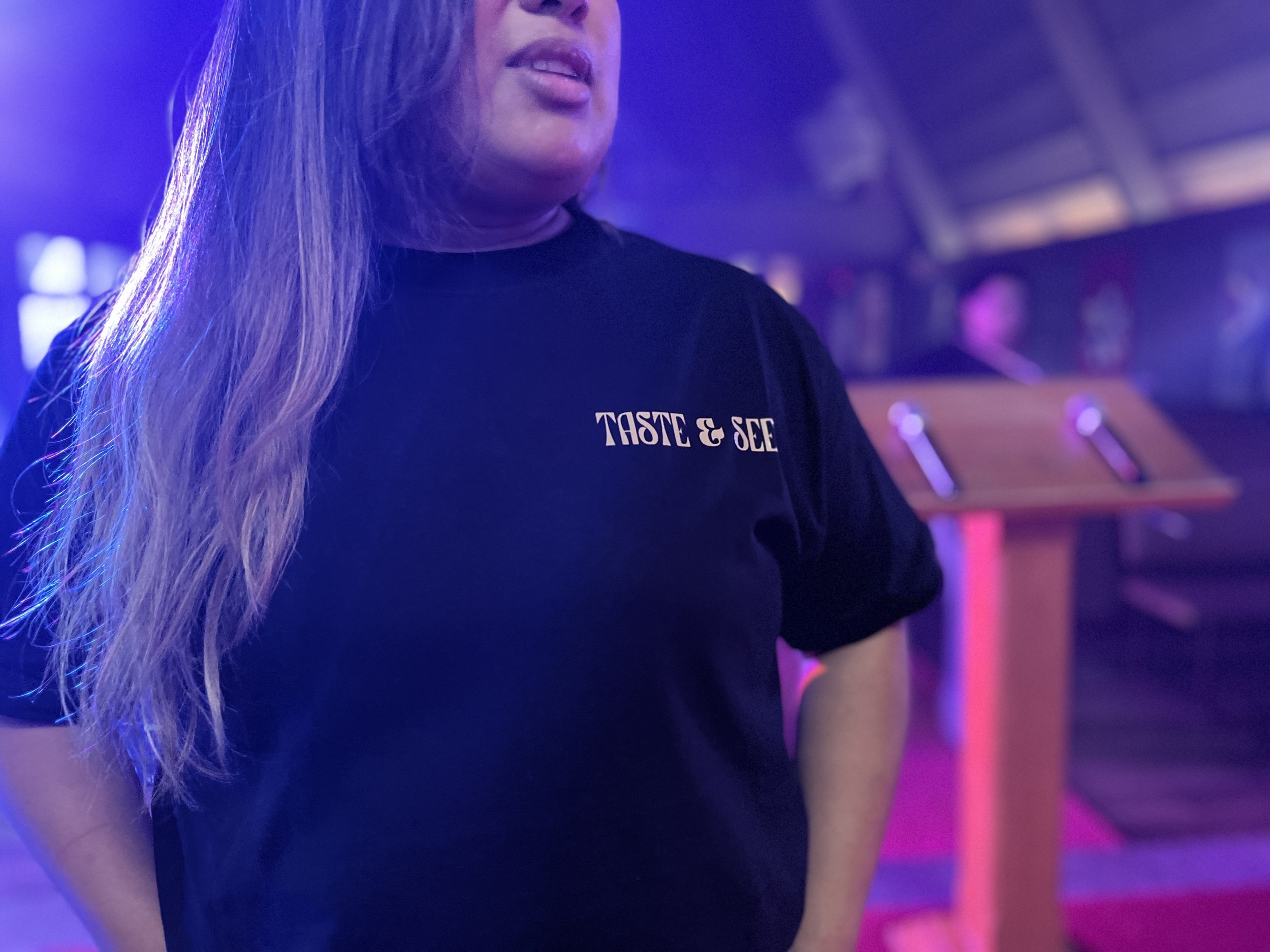 Close-up of a woman with long blonde hair, wearing a black T-shirt with the words 'TASTE & SEE' printed on it, in a dimly lit environment with purple and pink lights.