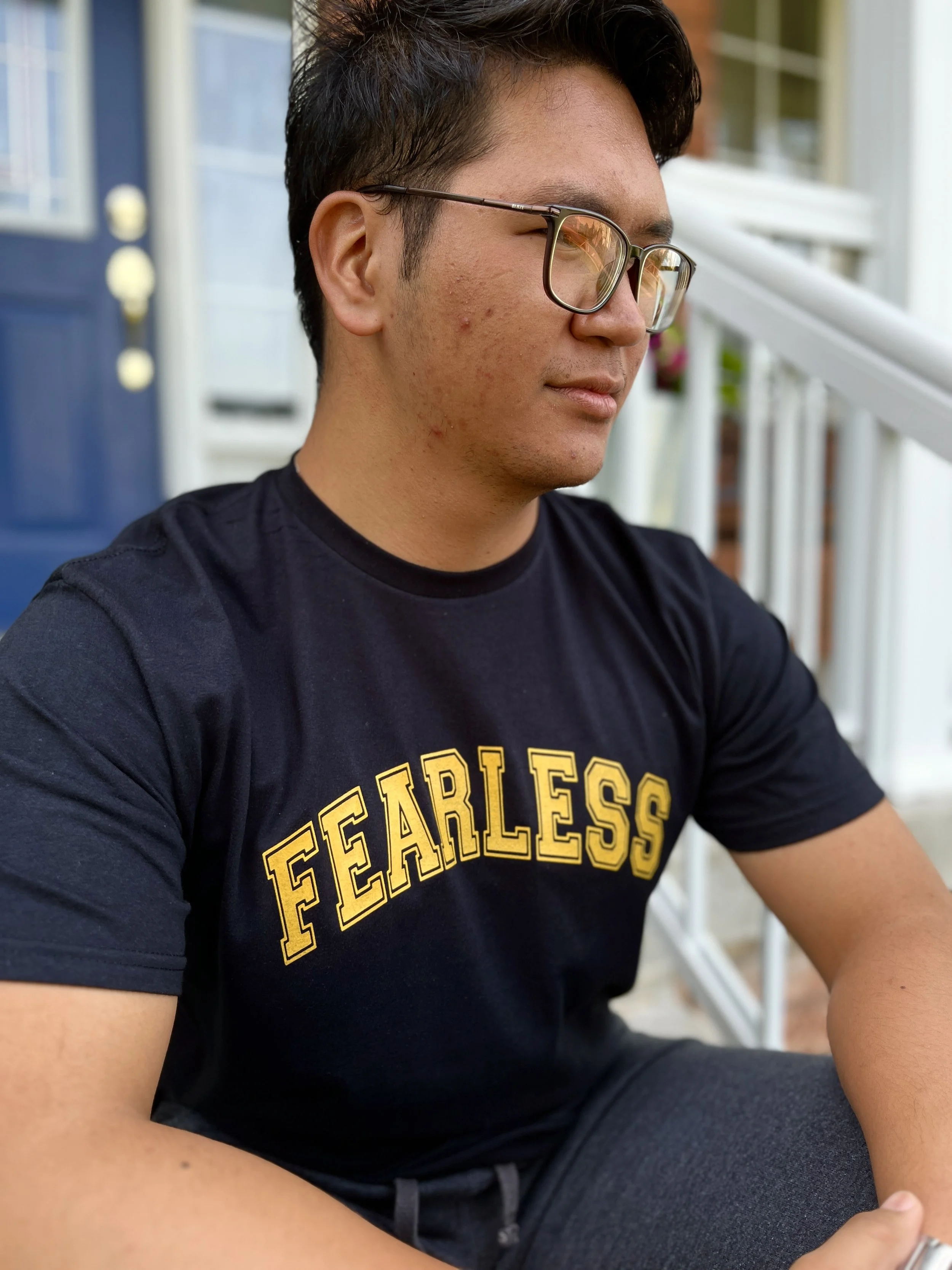 Close-up of a young man's face and upper body, wearing glasses and a black T-shirt with yellow text that says 'FEARLESS'. He appears to be sitting outside near a house with white siding and a blue door, with a porch or patio visible in the background