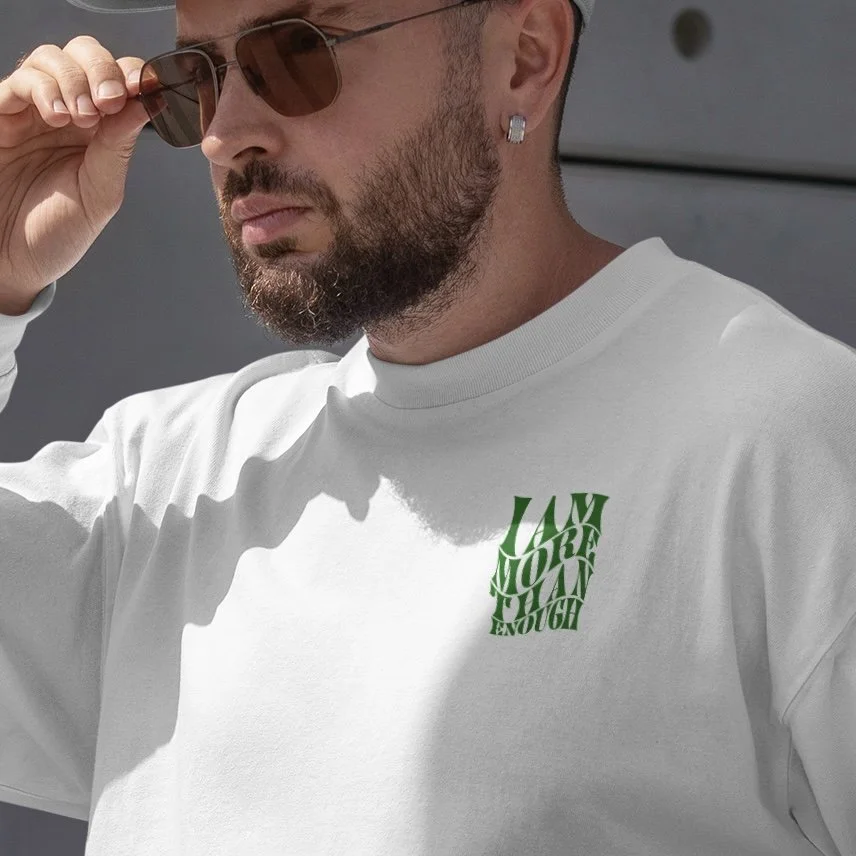 A man wearing sunglasses, a diamond stud earring, and a white T-shirt with green text that reads "I AM MORE THAN ENOUGH."