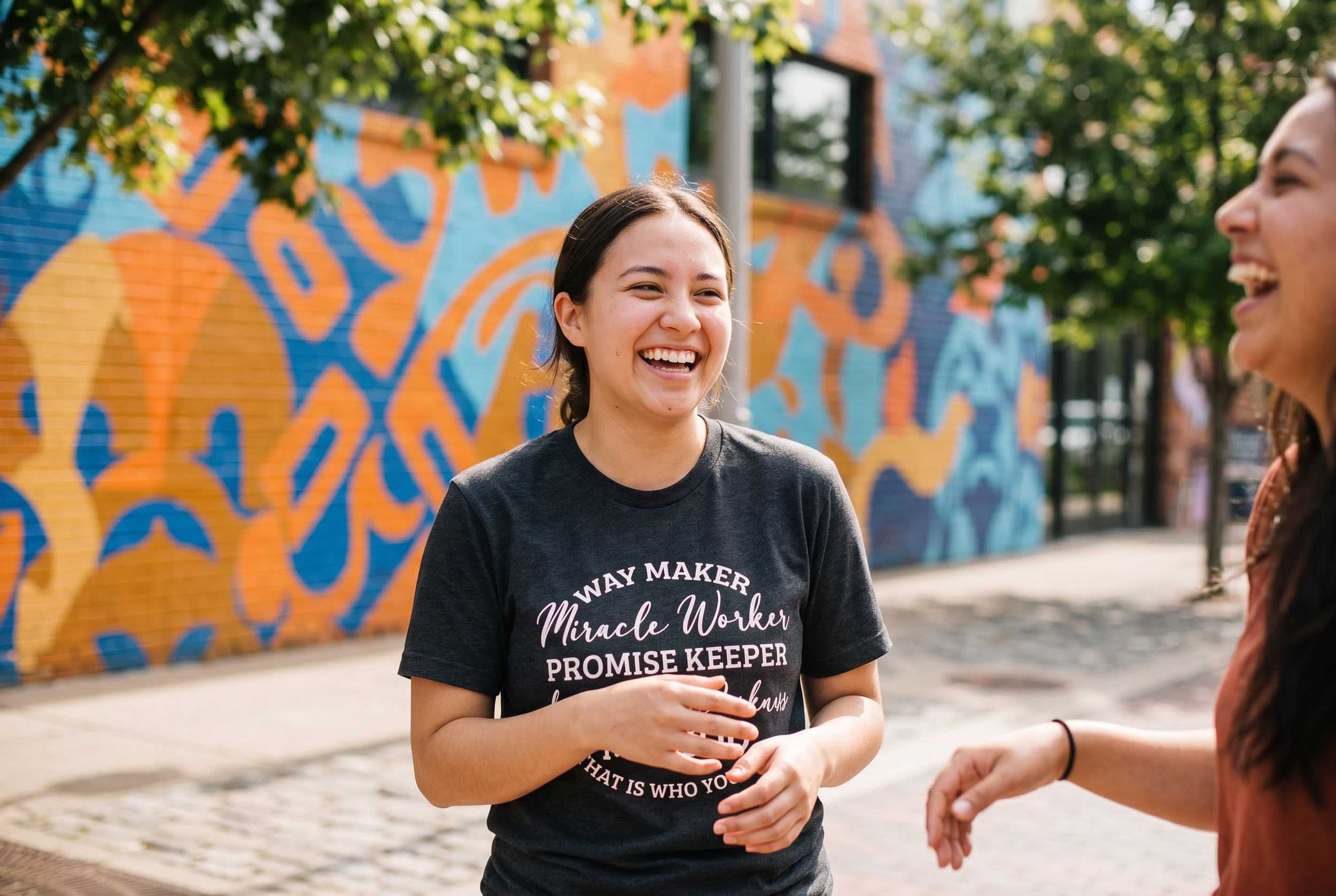 Two women smiling and talking outdoors, with a colorful mural in the background and trees providing shade.