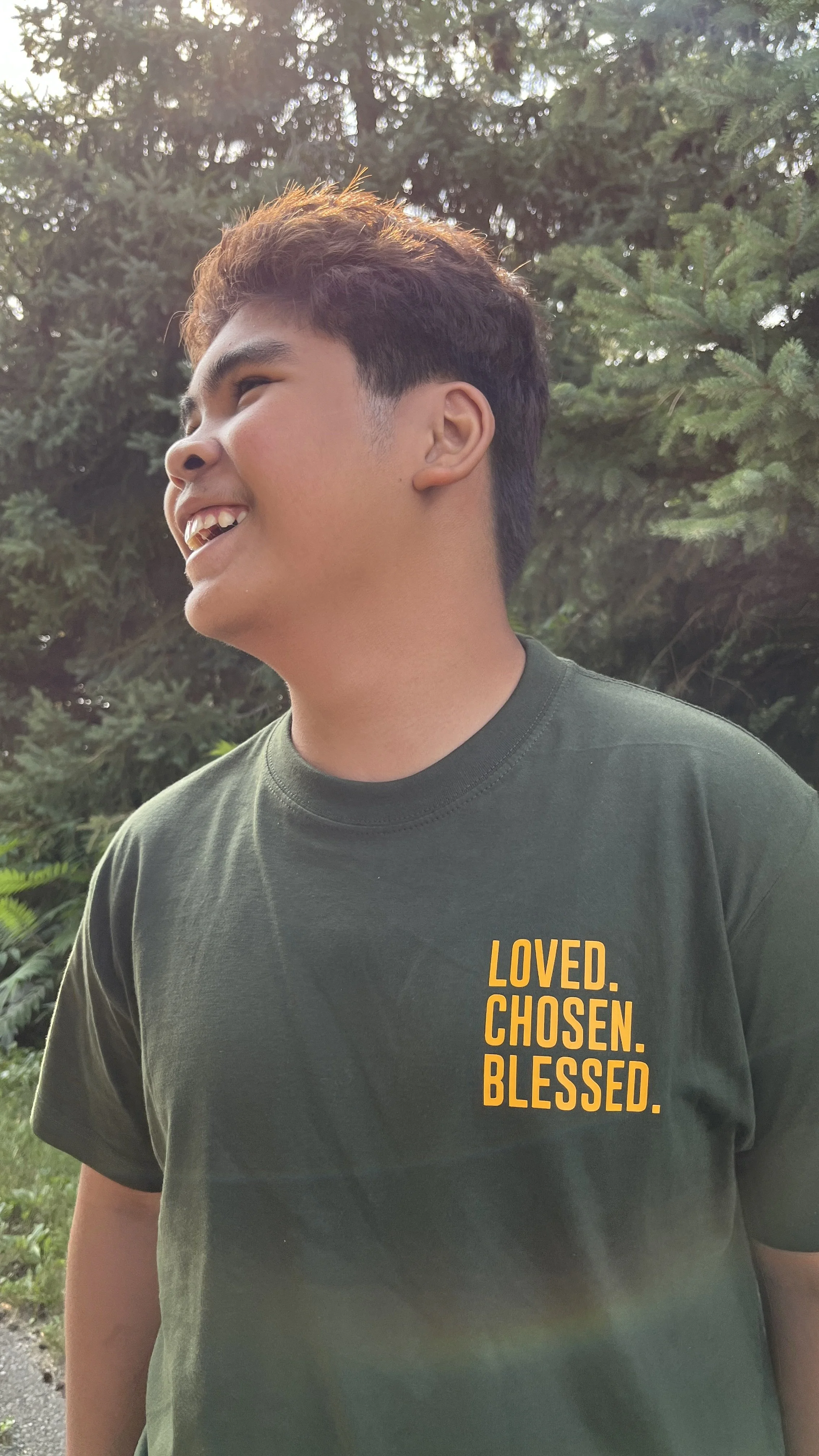 A young man laughing outdoors with trees in the background, wearing a dark green t-shirt with orange text that reads 'LOVED. CHOSEN. BLESSED.'