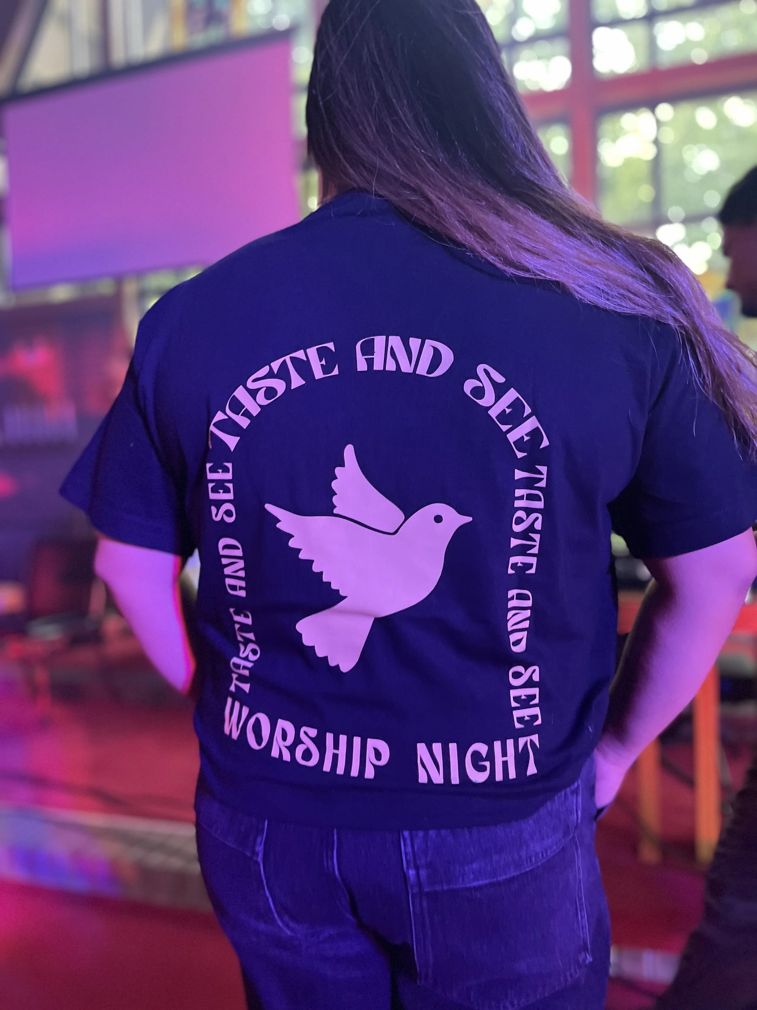 Person wearing a black T-shirt with a white dove graphic and the words 'WORSHIP NIGHT' on the back, standing in a dimly lit room with colorful lighting.