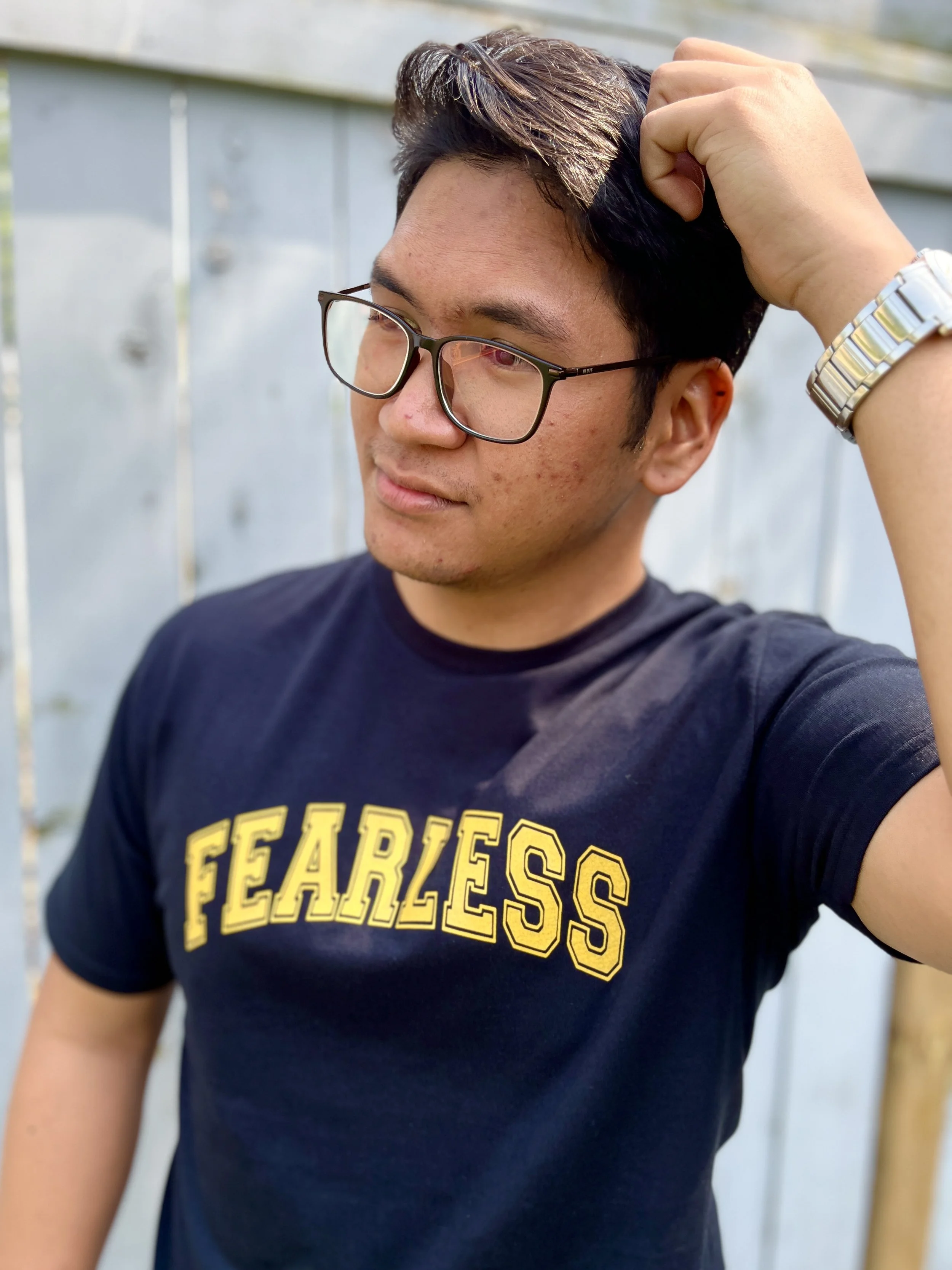 A young man with glasses and short dark hair, wearing a black t-shirt with yellow text, standing outdoors near a gray fence, touching his hair with his right hand.