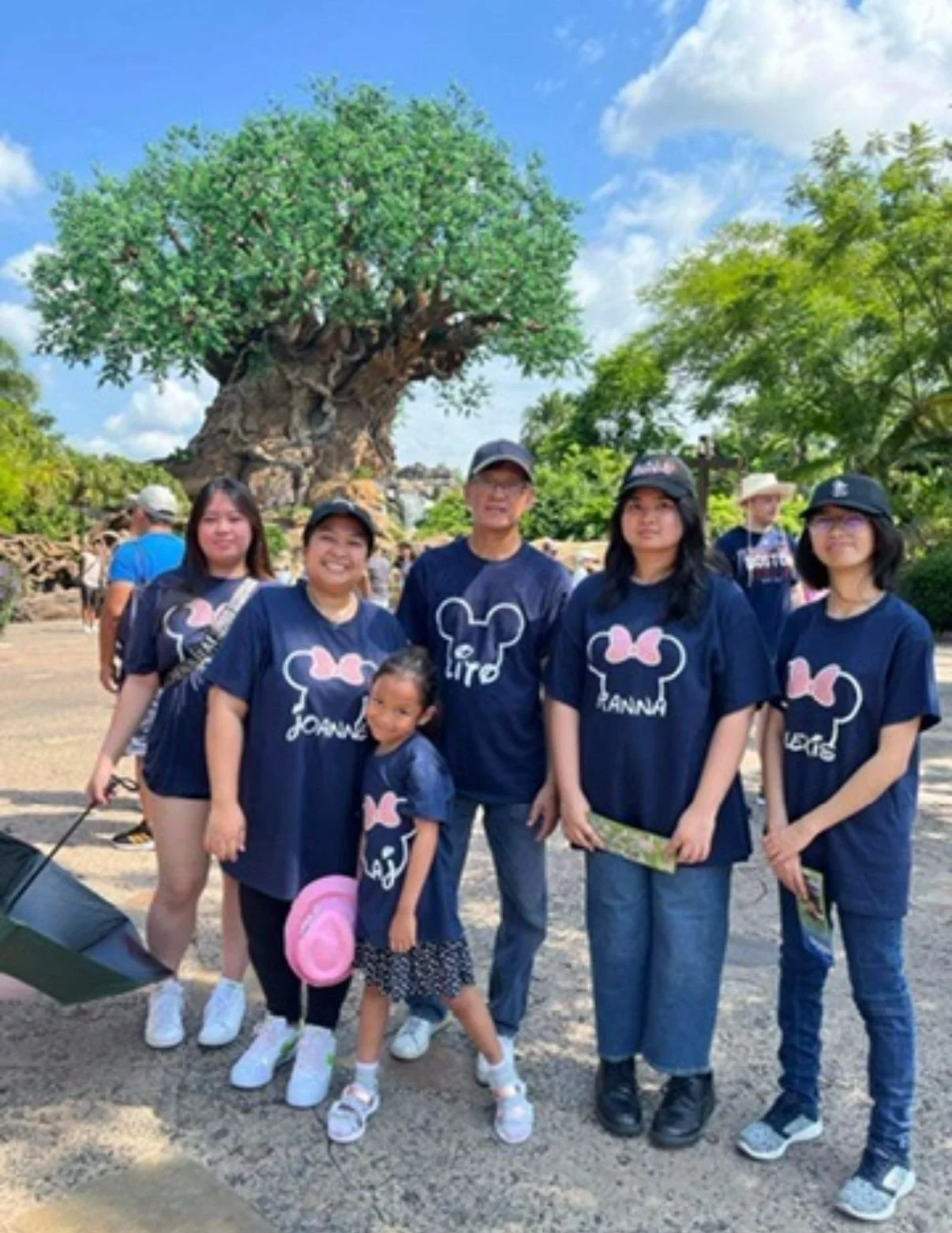 Group of six people, including children, posing in front of the Tree of Life at Disney's Animal Kingdom, wearing matching navy blue shirts with pink and white mouse ears and text.