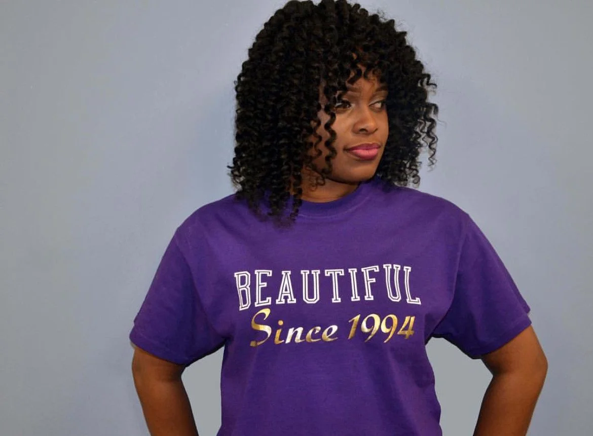 Woman with curly hair wearing a purple T-shirt that says 'BEAUTIFUL Since 1994' standing against a gray wall.