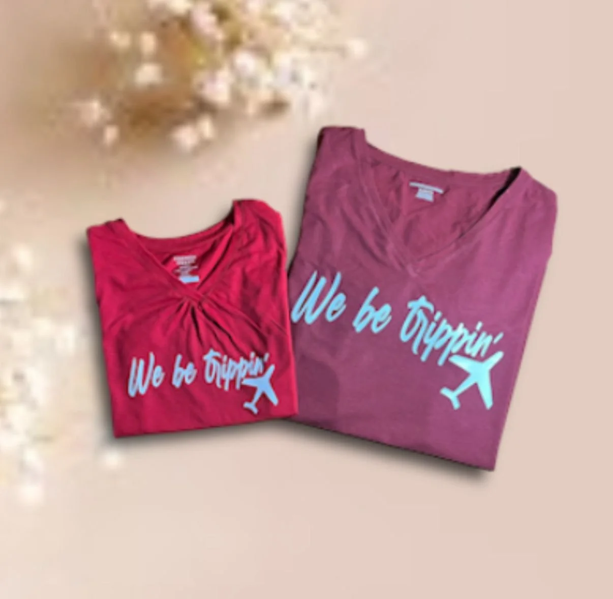 Two folded pink T-shirts with the phrase "We be Trippin'" and an airplane graphic, lying on a light-colored surface near some white flowers.
