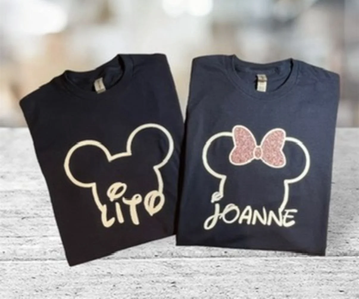 Two navy blue T-shirts with Mickey Mouse head outlines and personalized names, one reads 'LITO' with a mouse face, the other 'JOANNE' with a mouse face and pink bow.