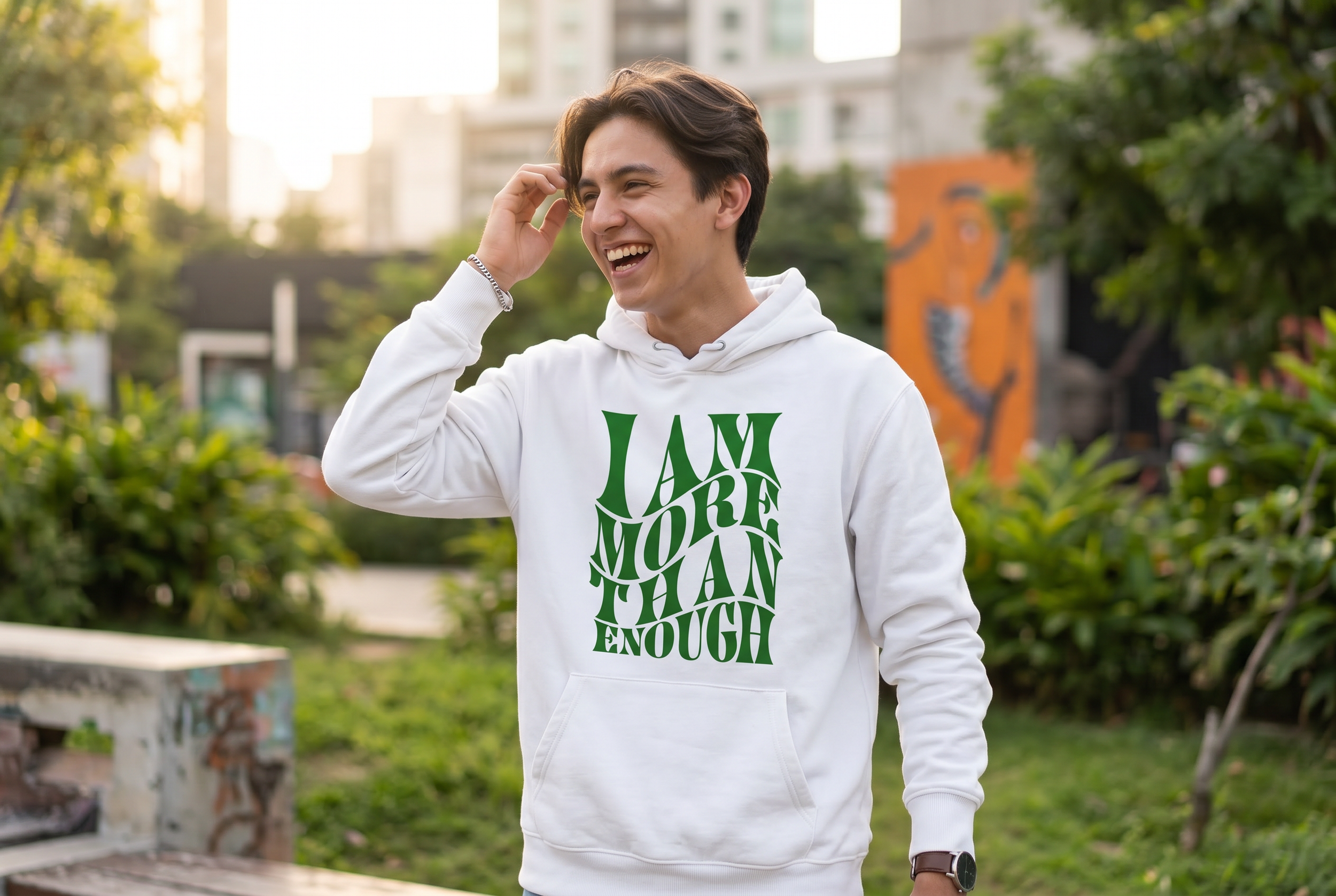A young man with dark hair smiling and wearing a white hoodie with green text, standing outdoors in a park with greenery and buildings in the background.