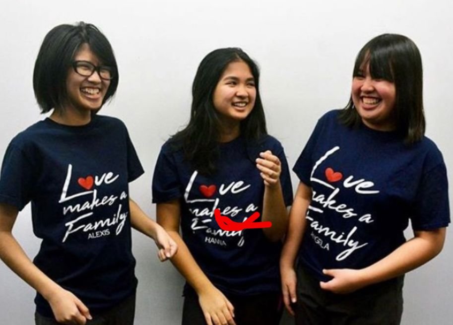 Three young women wearing matching navy blue T-shirts with the phrase 'Love makes a Family' and their names, standing against a plain white wall, smiling and laughing together.