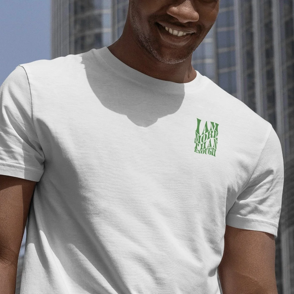 Man wearing a white T-shirt with a small green graphic that reads "I AM MORE THAN ENOUGH" on the chest side, outdoors with tall buildings in the background.