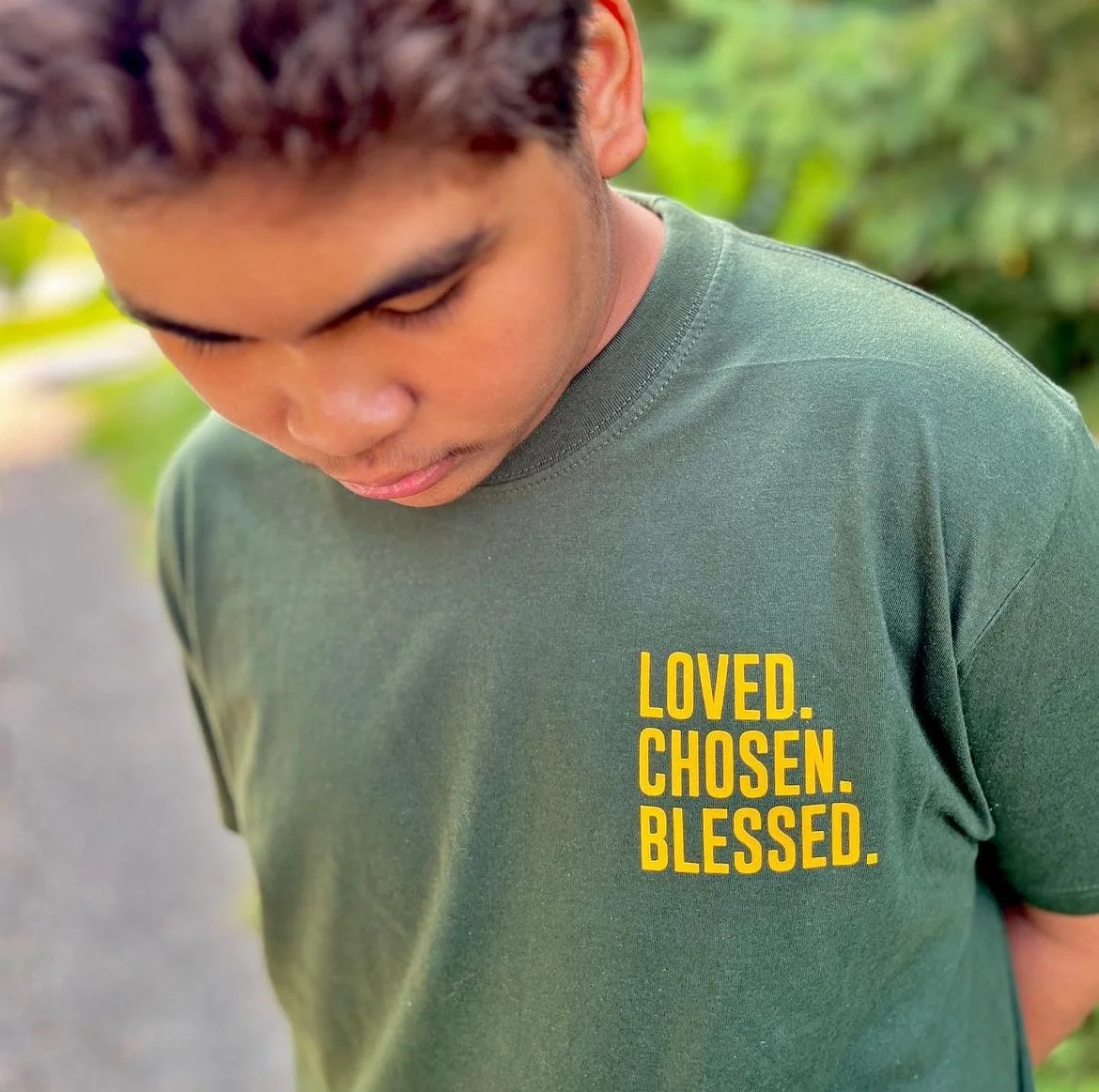 Close-up of a young man wearing a green t-shirt with yellow text that reads "LOVED. CHOSEN. BLESSED." outdoors with green foliage in the background.