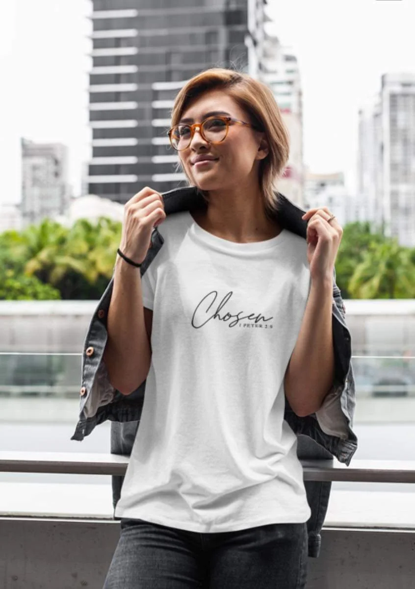A woman with short hair and sunglasses standing outdoors on a balcony with a cityscape of tall buildings in the background. She is wearing a white T-shirt with the word "Chosen" and a black jacket draped over her shoulders.