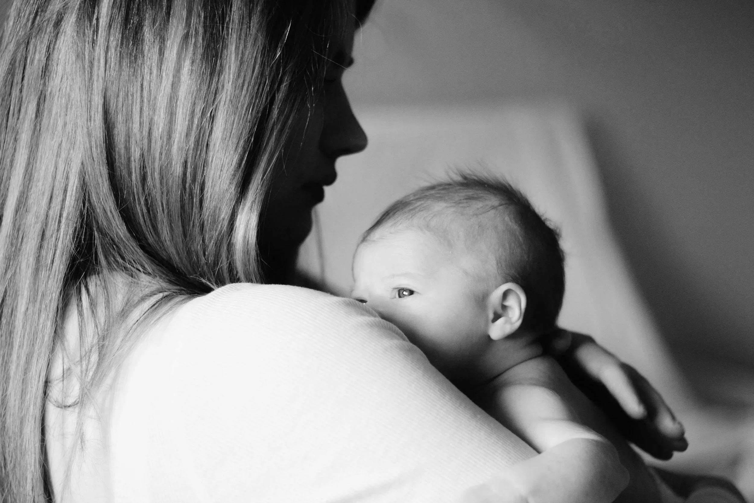 A black and white photograph of a woman holding a young child close to her chest. The woman has long hair and is looking down, while the child's face is partially visible as he rests against her shoulder.