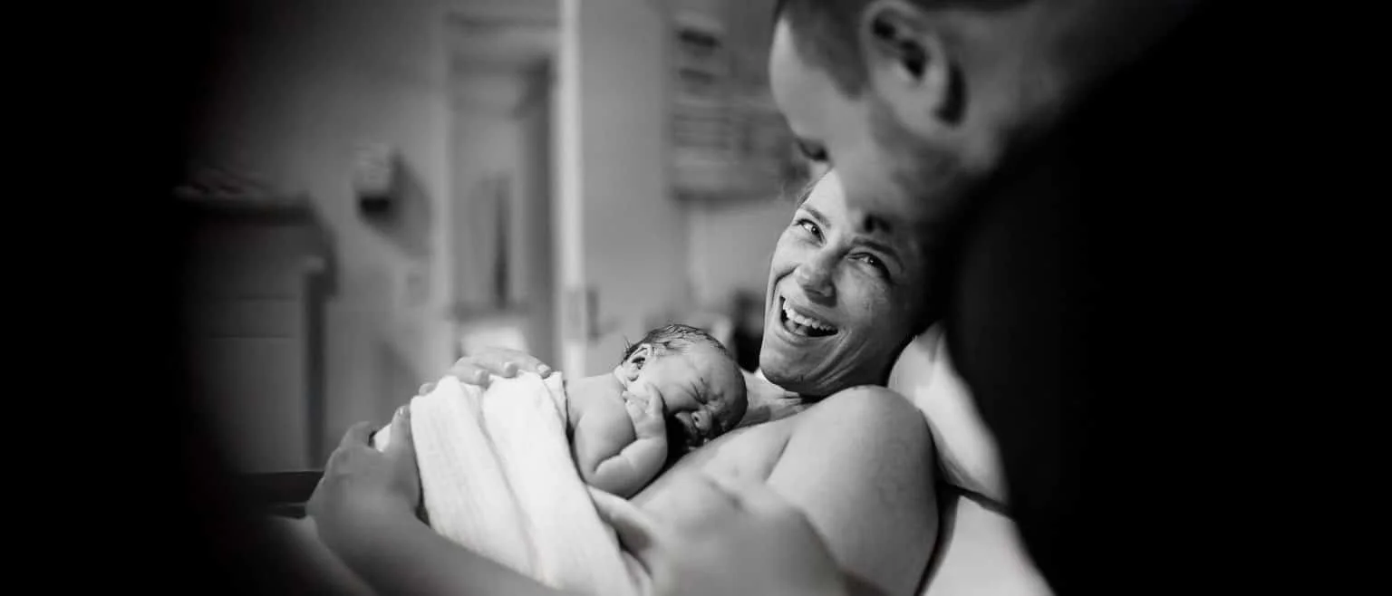 A newborn baby resting on a woman's chest, with her giving a joyful smile, in a hospital room.