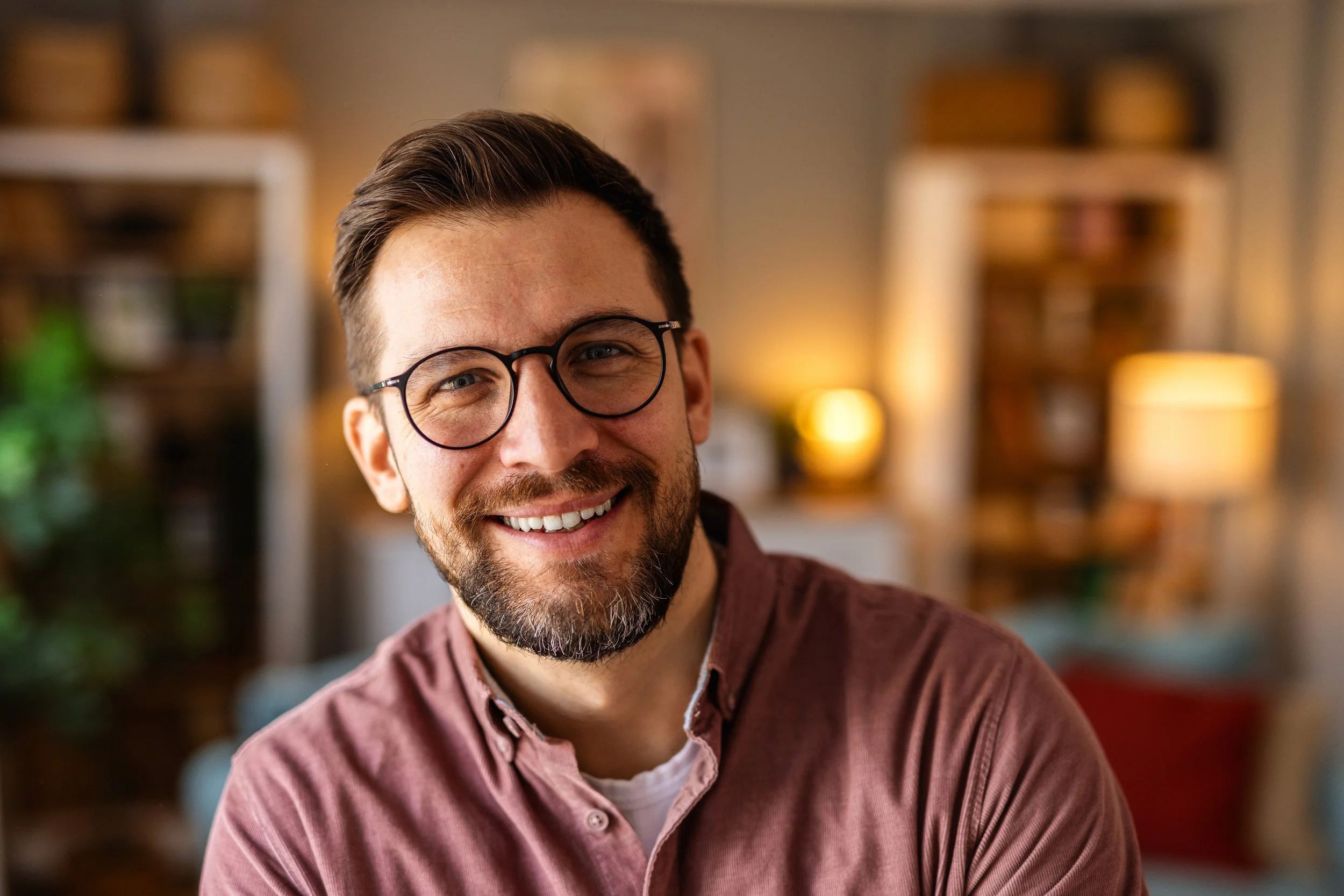 A smiling man with glasses and a beard, wearing a maroon shirt, in a cozy room with warm lighting.