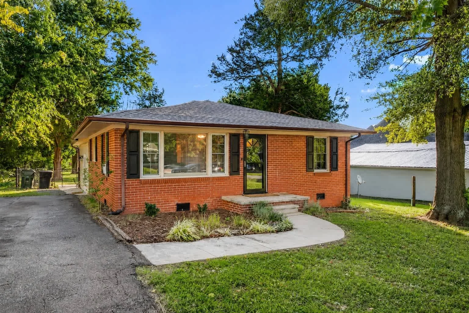 A single-story brick house with black shutters and front steps, surrounded by green trees and a well-maintained lawn, during daytime.