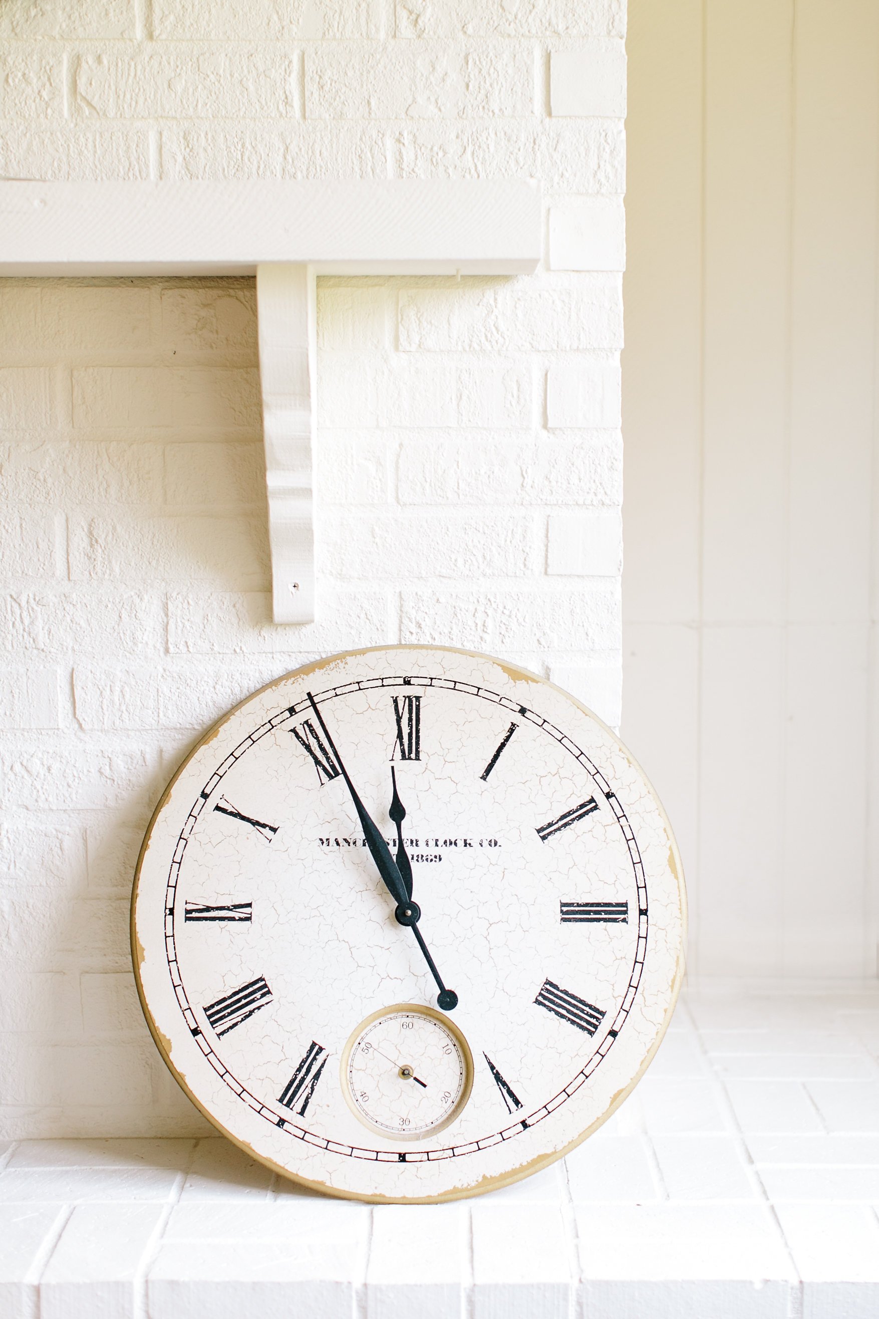 White vintage clock with Roman numerals and a small second subdial, mounted on a white brick wall in a room with white panels.