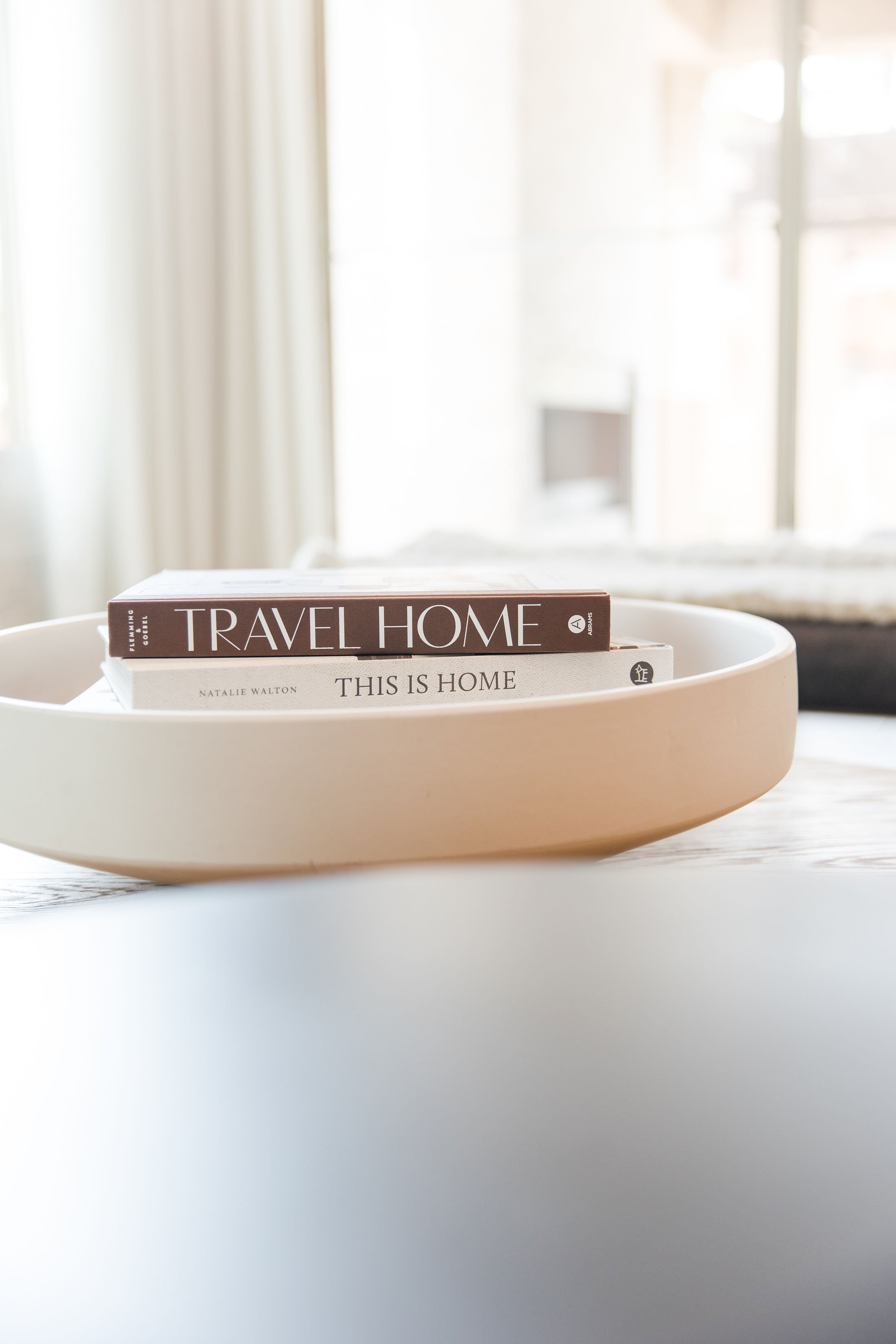 Two books titled "Travel Home" by Flemming Goell and "This Is Home" by Natalie Walton placed in a white ceramic bowl on a white surface, with a bright, softly blurred window background.