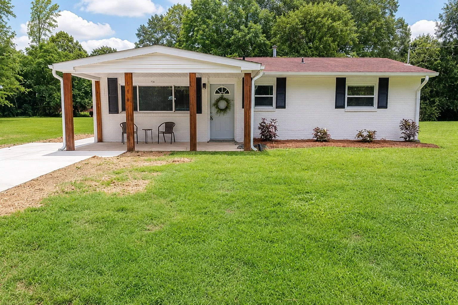 Front view of a white brick house with a red roof, black shutters, and a small covered porch with wooden posts. There are two chairs and a small table on the porch, with a wreath on the front door. The lawn is green with some freshly tilled soil and a few small bushes in front of the house. Tall trees are visible in the background under a partly cloudy sky.