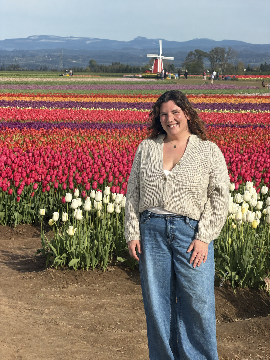 Woman smiling in a beige cardigan and blue jeans standing in a colorful tulip field with a traditional windmill in the background and mountains under a partly cloudy sky.