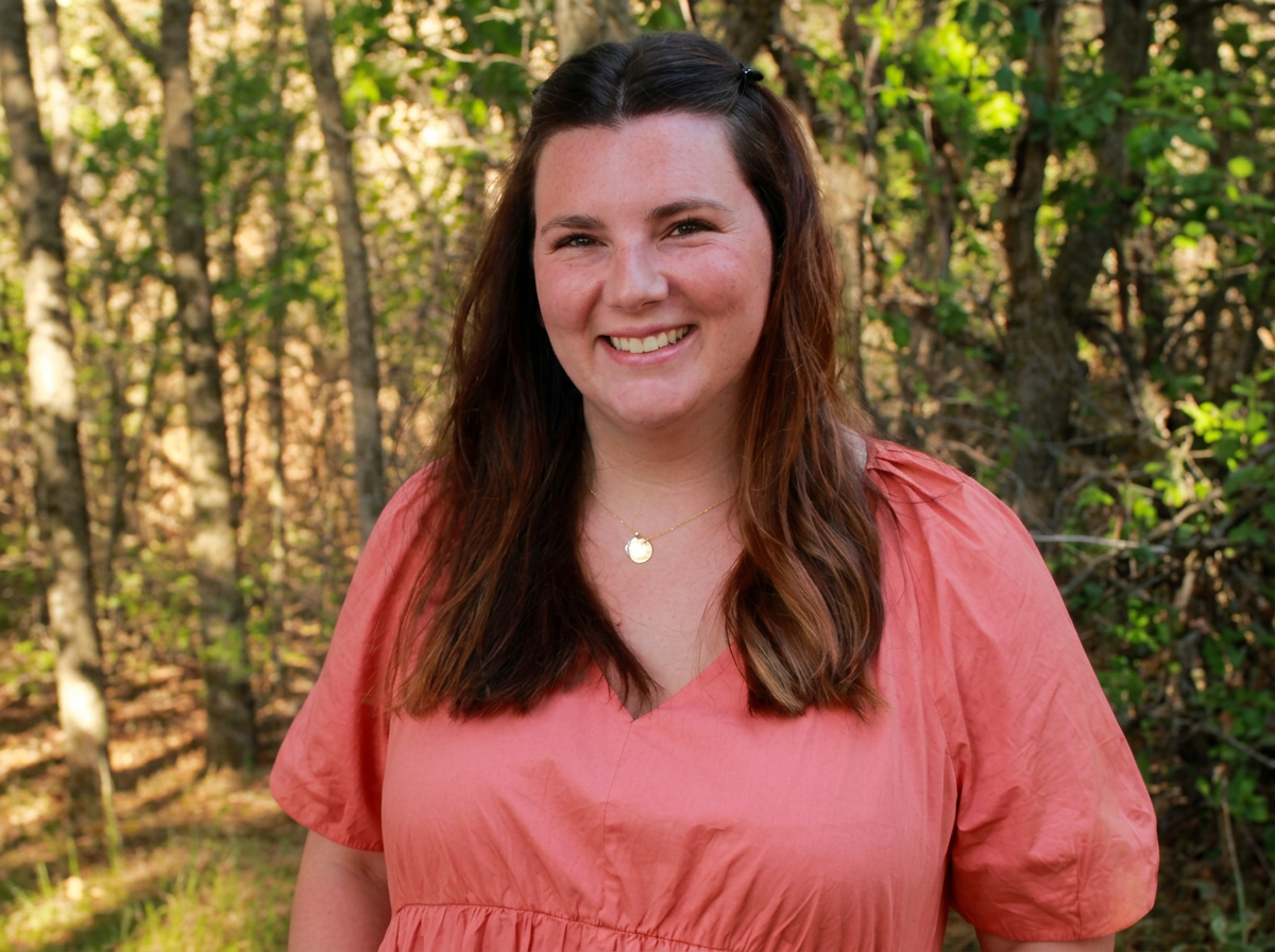 Portrait of a smiling woman with brown hair, wearing a coral-colored top and a gold necklace, standing outdoors in a forested area with green leaves and trees in the background.