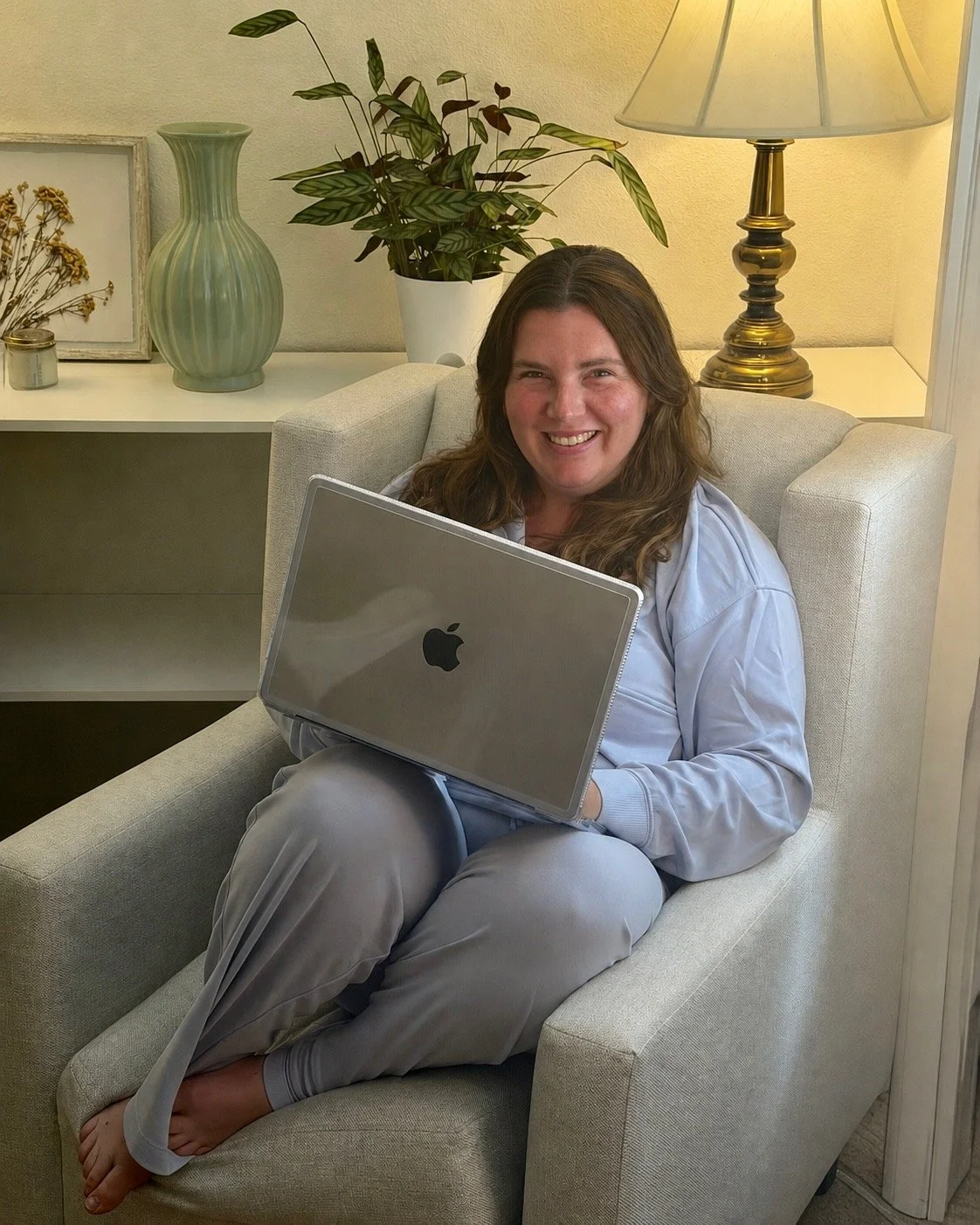 A woman with long brown hair sitting in a beige armchair, smiling, holding a silver MacBook laptop on her lap indoors near a side table with decorative items and a lit lamp.
