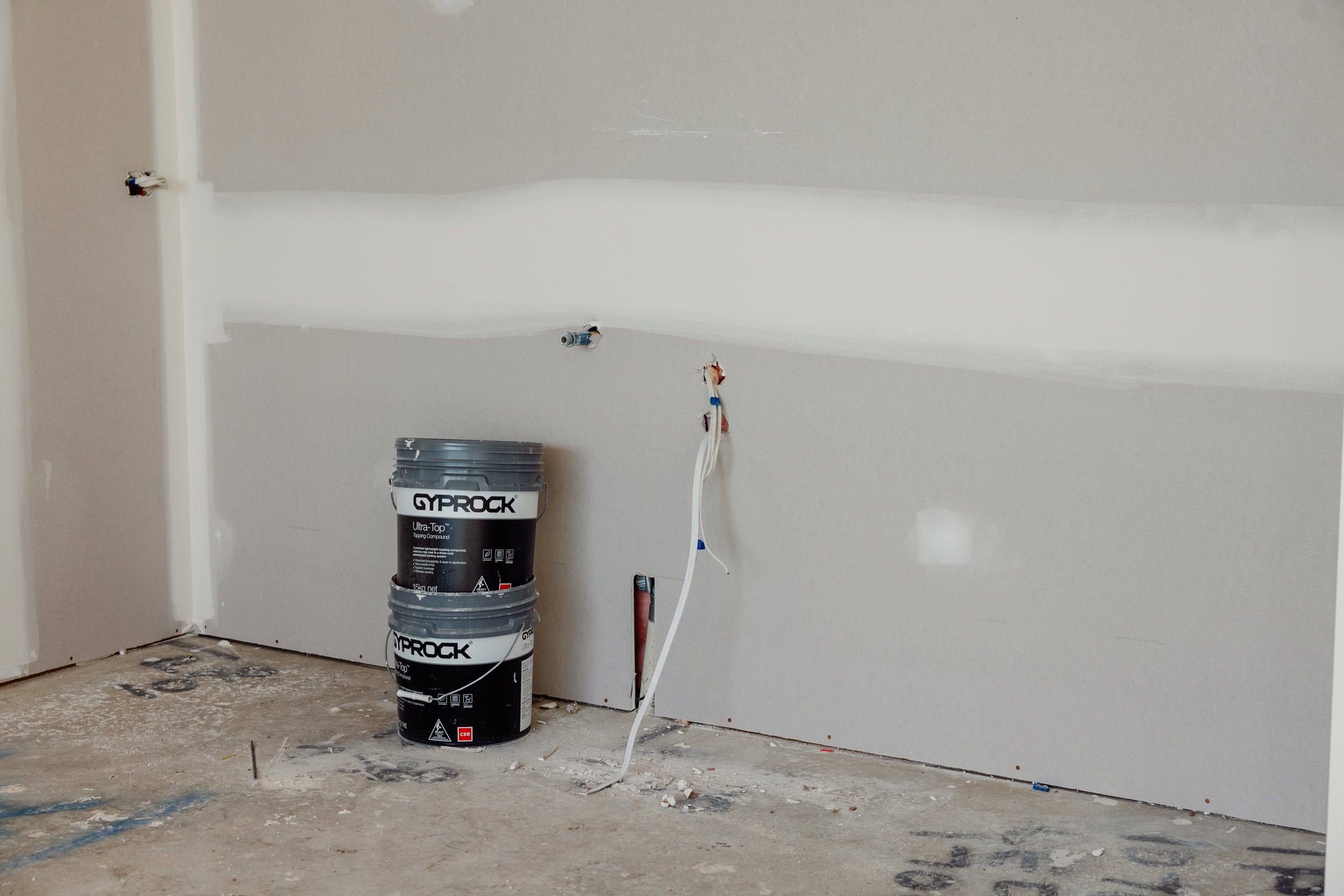 Interior wall under construction with two stacked buckets of drywall compound, exposed wiring, and unfinished floor.