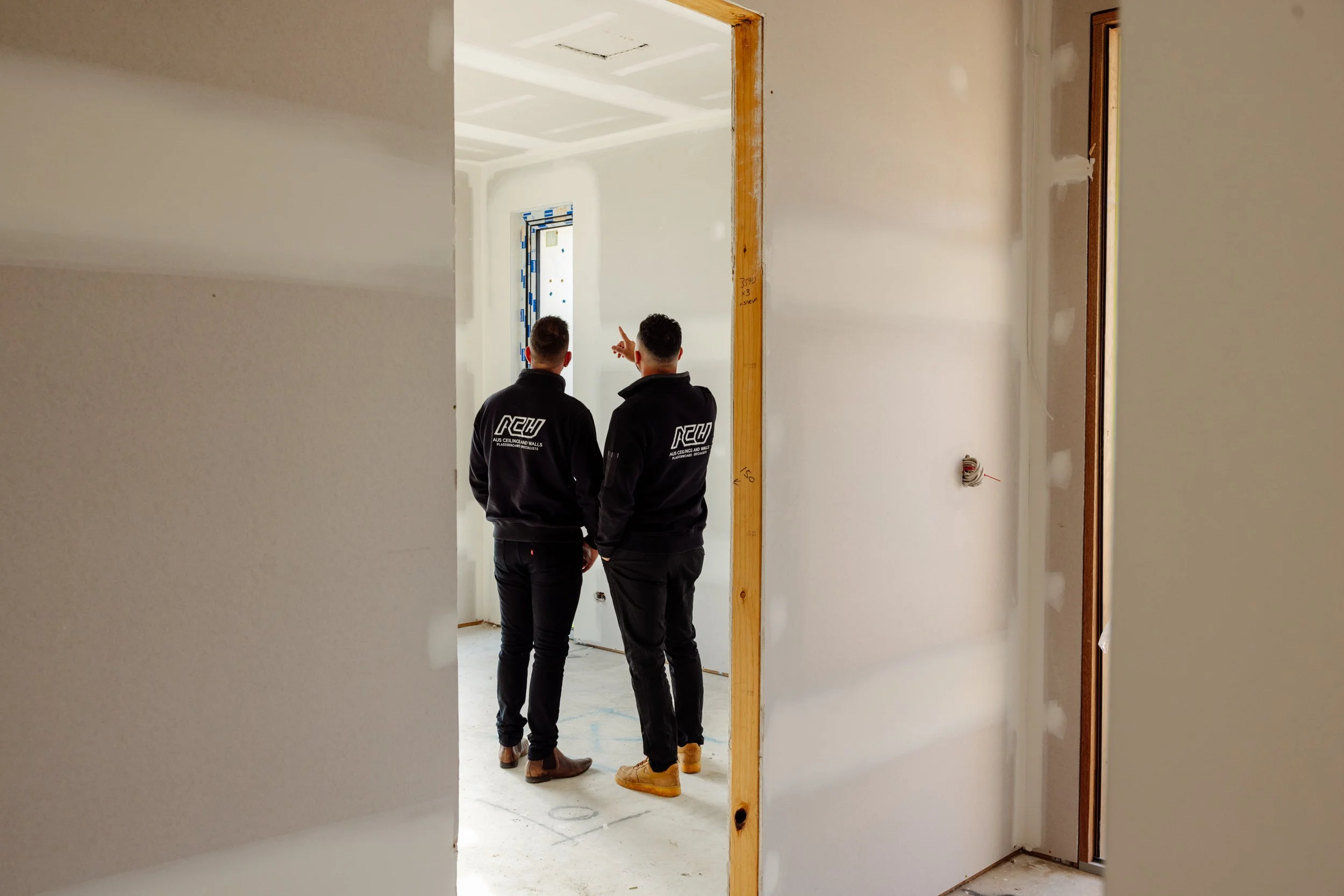 Two men wearing black hoodies standing in doorway of a room under construction, pointing towards the interior.