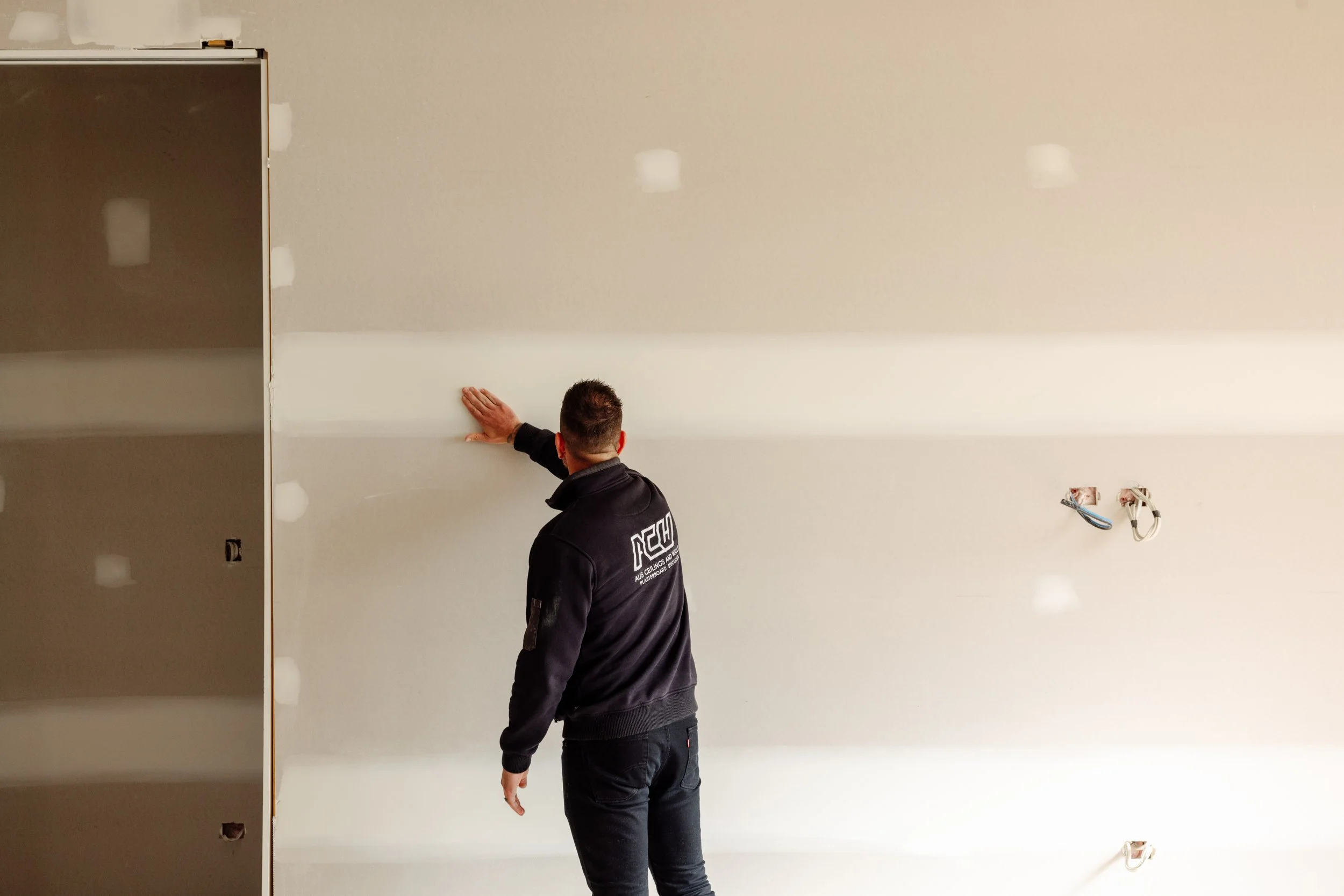 A person wearing a black jacket with white text on the back is standing in front of a wall, working on drywall installation or repair. The room appears to be under construction or renovation, with visible electrical wiring and uninstalled fixtures.