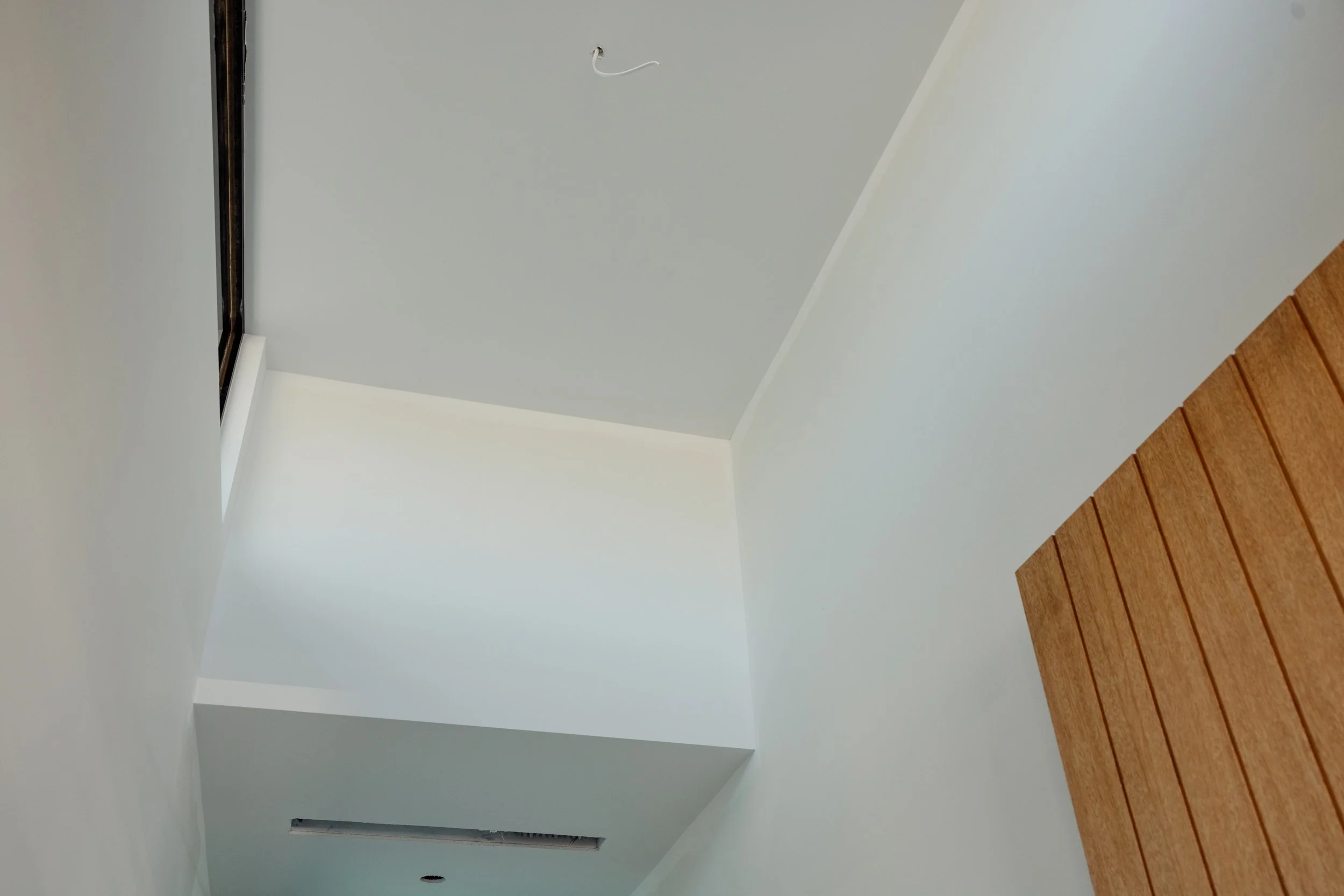 Ceiling with a small hole and a wire hanging down, corner of a room with white walls, wood paneling on one wall, and a vent opening near the ceiling.