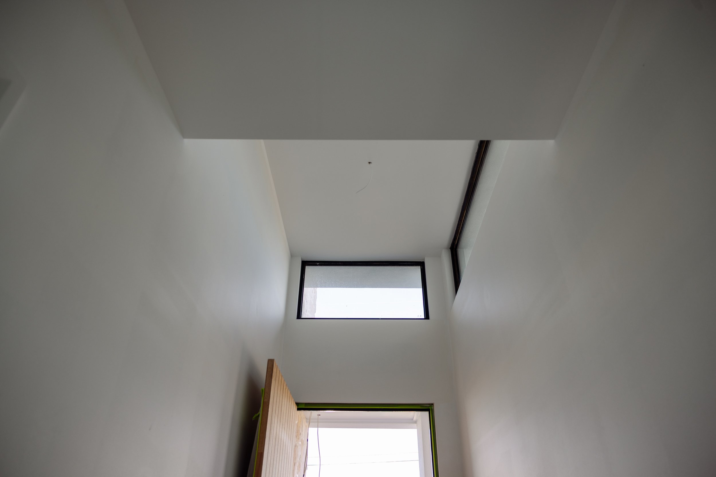 Empty room with white walls, high ceiling, large windows at the far end letting in natural light, and a small wooden piece of furniture on the left side near the wall.