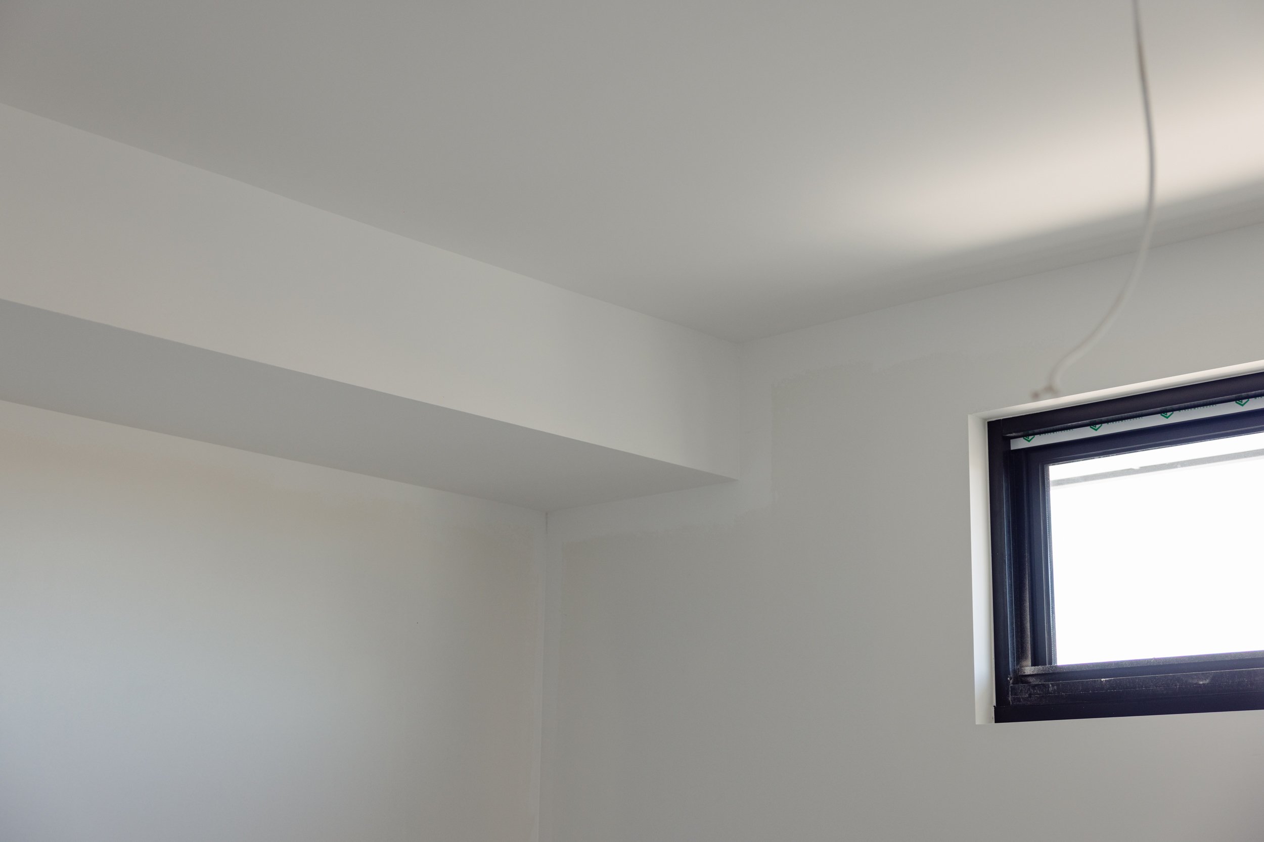 Part of a white ceiling and wall with a black-framed window in the corner, showing a partially open venetian blind, and a white electrical wire near the ceiling.