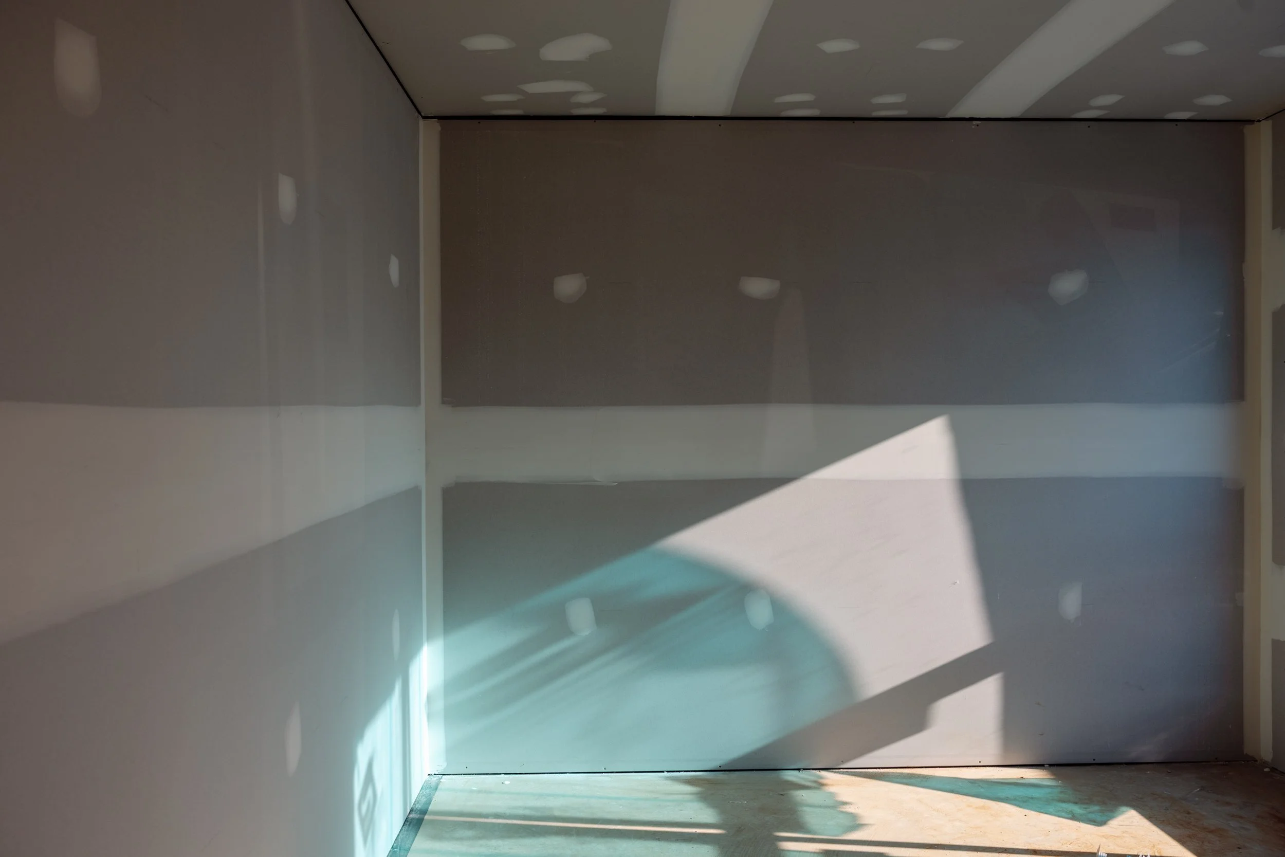 An empty unfinished room with gray drywall and shadows cast on the walls and ceiling by a window.