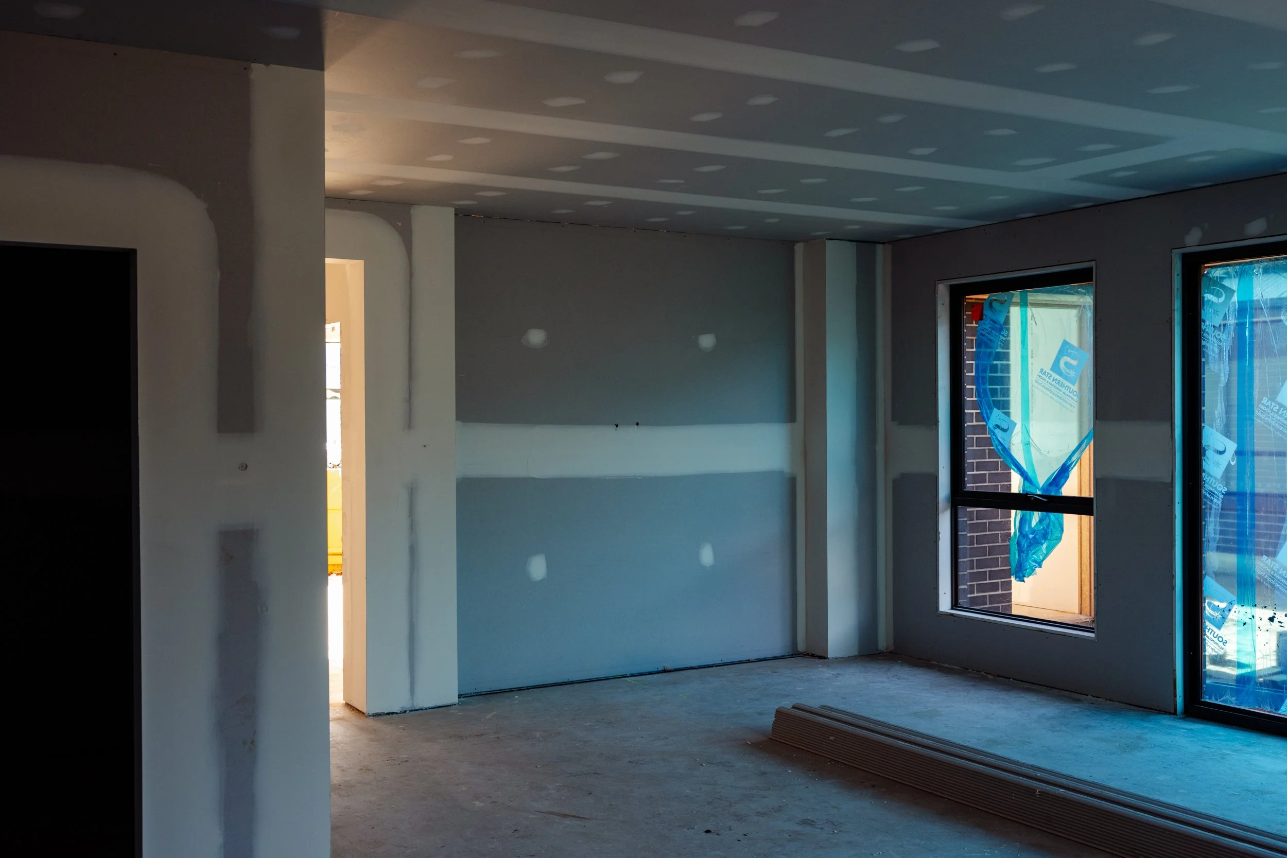 Interior of a room under construction with unfinished walls, a floor, and large windows wrapped in blue protective plastic, letting in natural light.