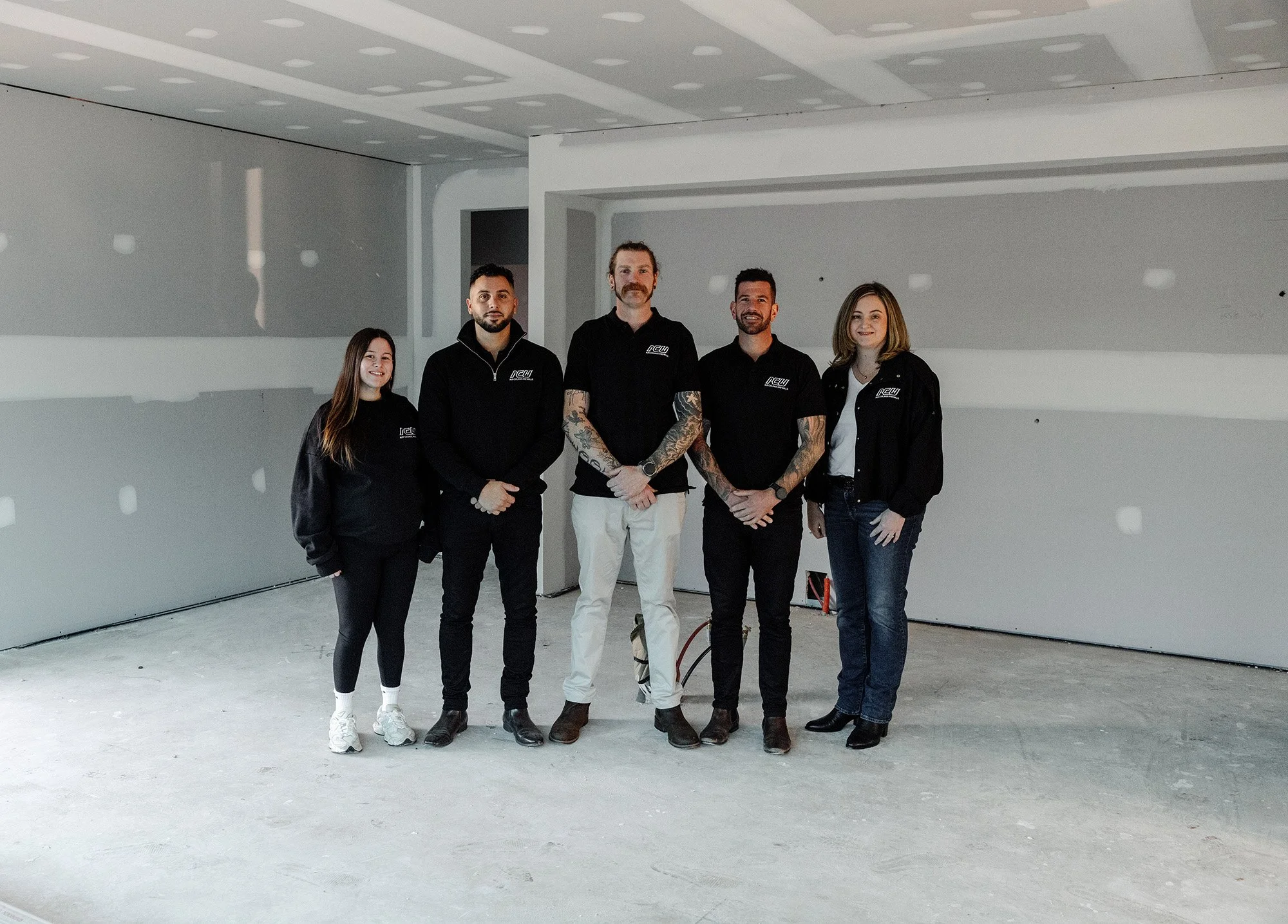 Group of five people standing in an unfinished room with drywall and no flooring.