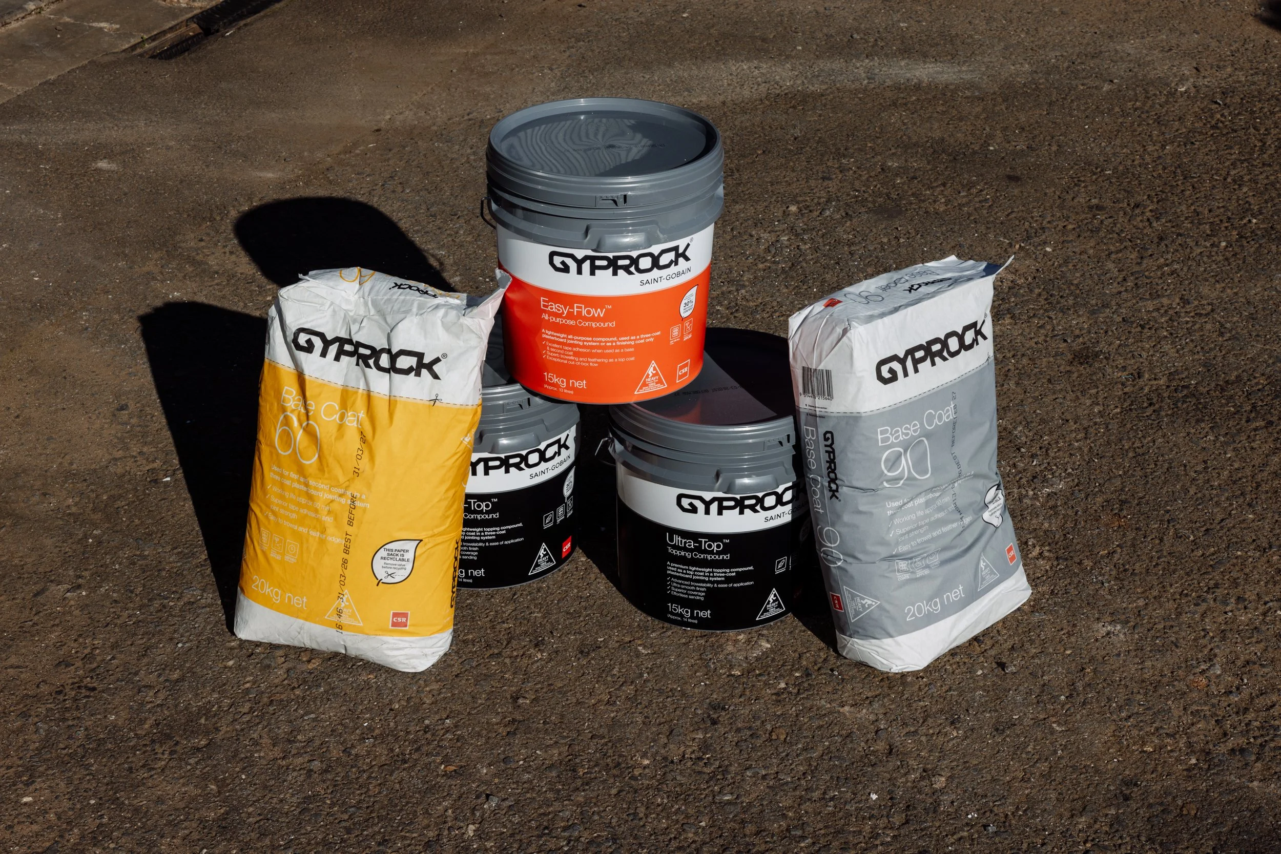 Stacked black and gray buckets with GYPROK branding, surrounded by bags of GYPROK base coat and other construction materials on a gravel surface.