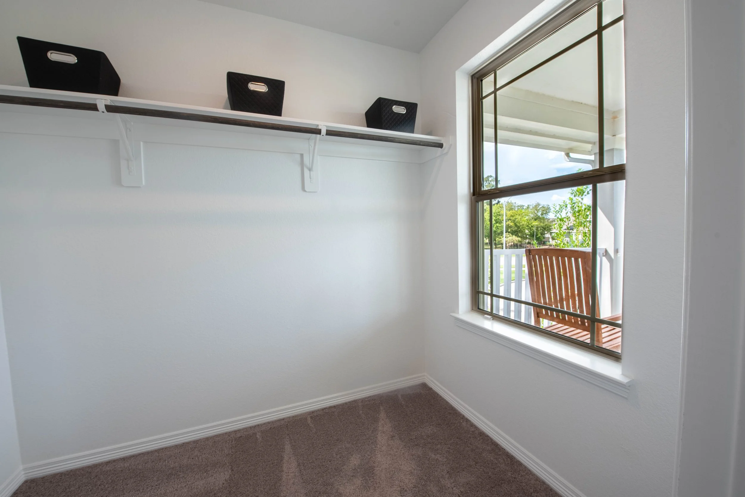 Empty room with carpet flooring, white walls, a large window with a view outside, and a white shelf attached to the wall with three black storage baskets on top.