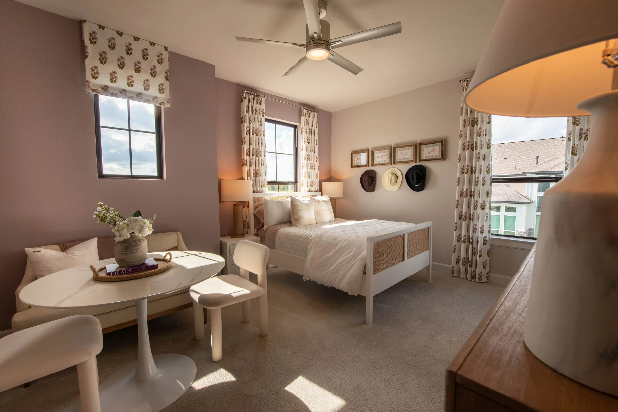 A cozy bedroom with a white bed, floral curtains, and framed hats on the wall. A small table with a vase of flowers and a tray with books is beside the bed. Two windows let in natural light, and a ceiling fan is above.