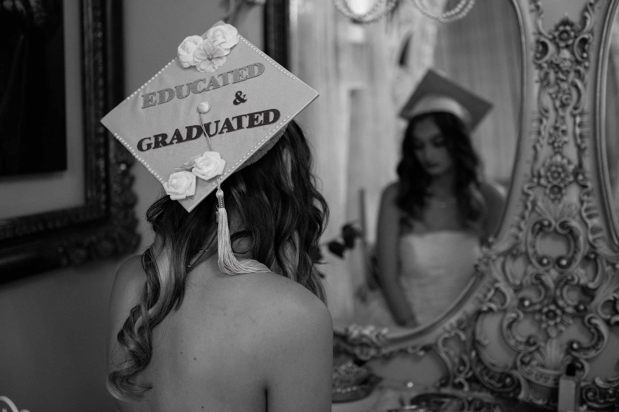 A graduate woman with long curled hair wearing a decorated cap, looking in a mirror in an ornate room.