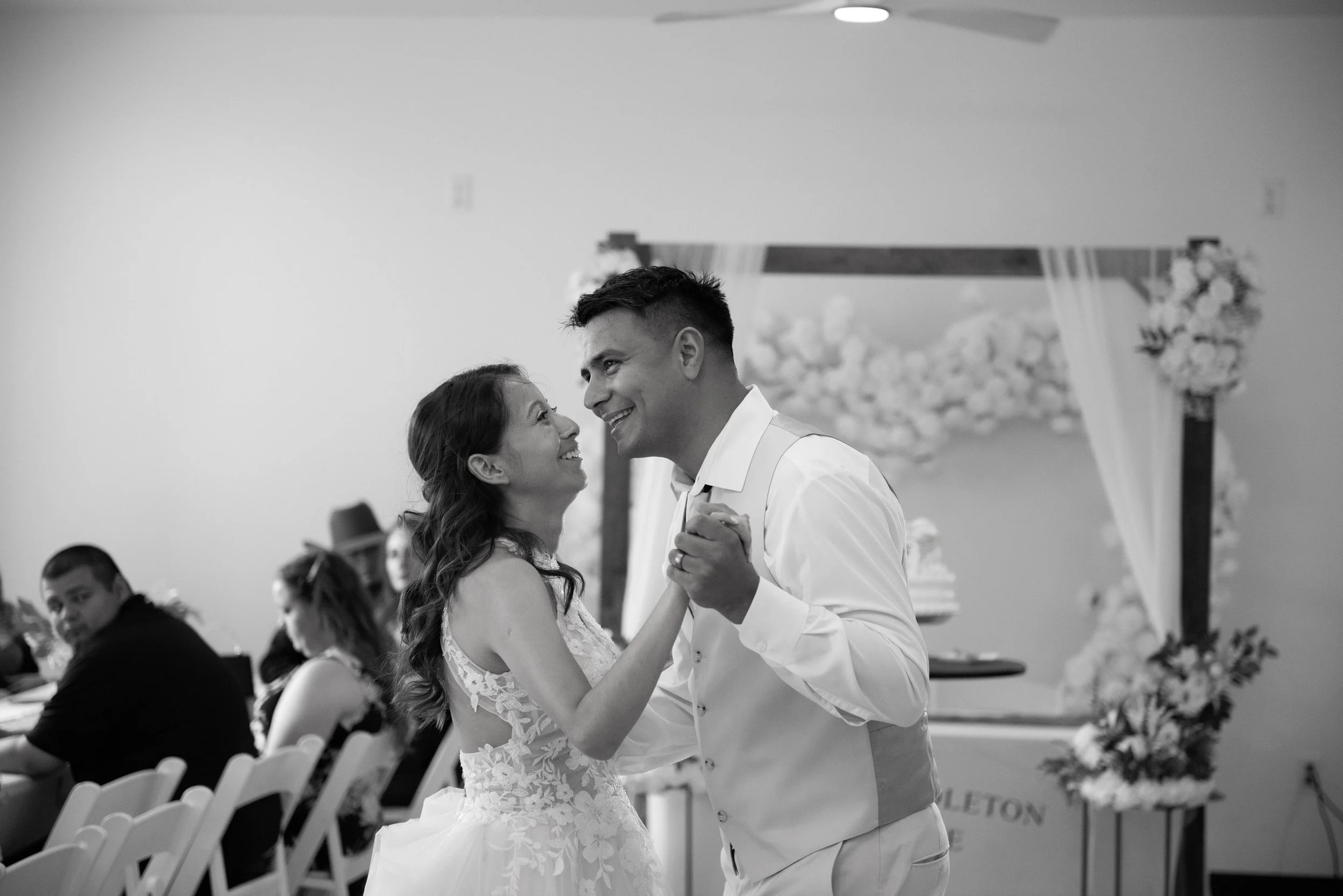 A couple dancing at their wedding reception. The bride and groom are smiling and looking at each other, surrounded by seated guests and decorative flowers in the background.