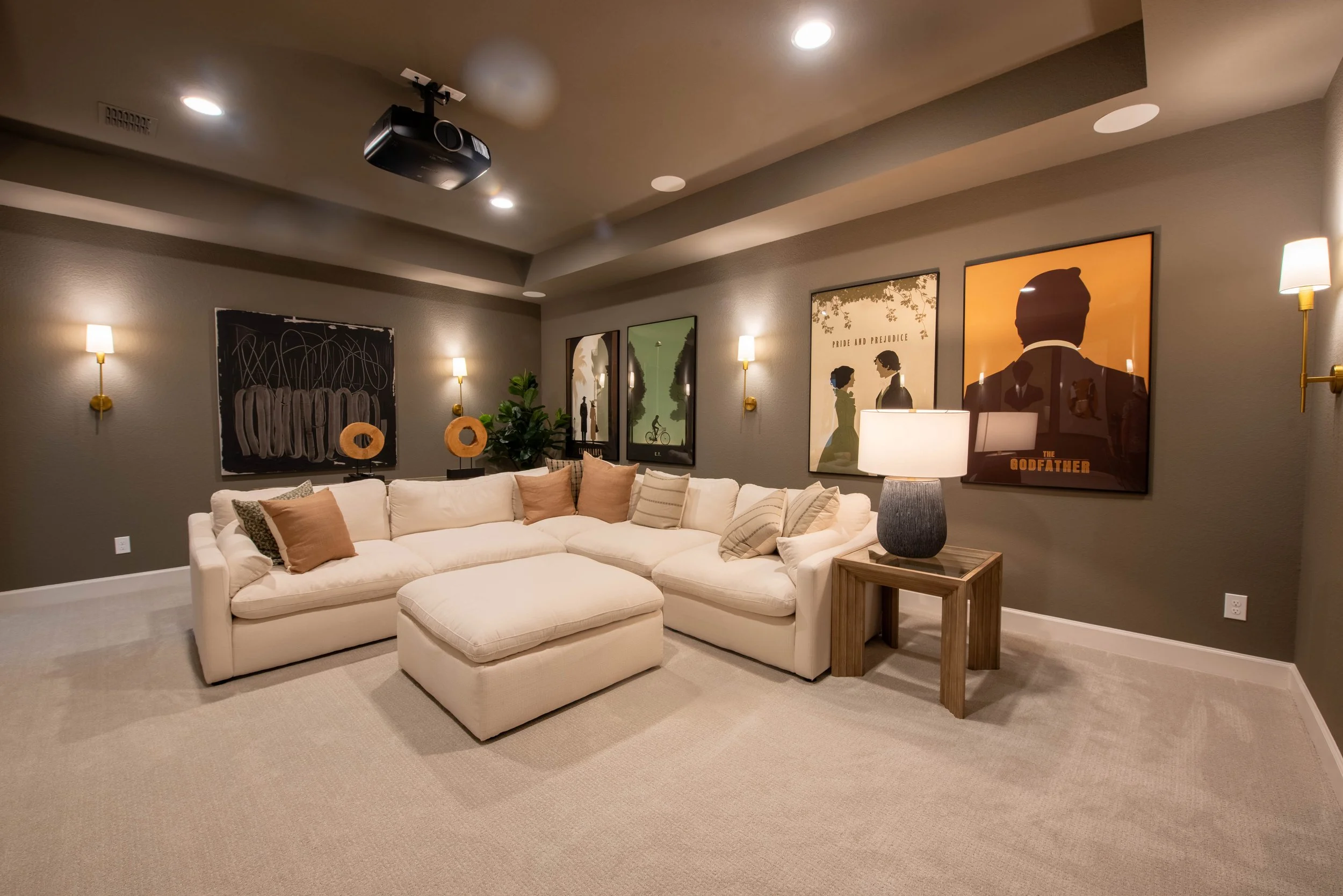 Living room with white sectional sofa, beige and neutral pillows, a wooden side table with lamp, framed artwork on the gray wall, beige carpet, with ceiling lights and wall sconces, and a ceiling-mounted projector.