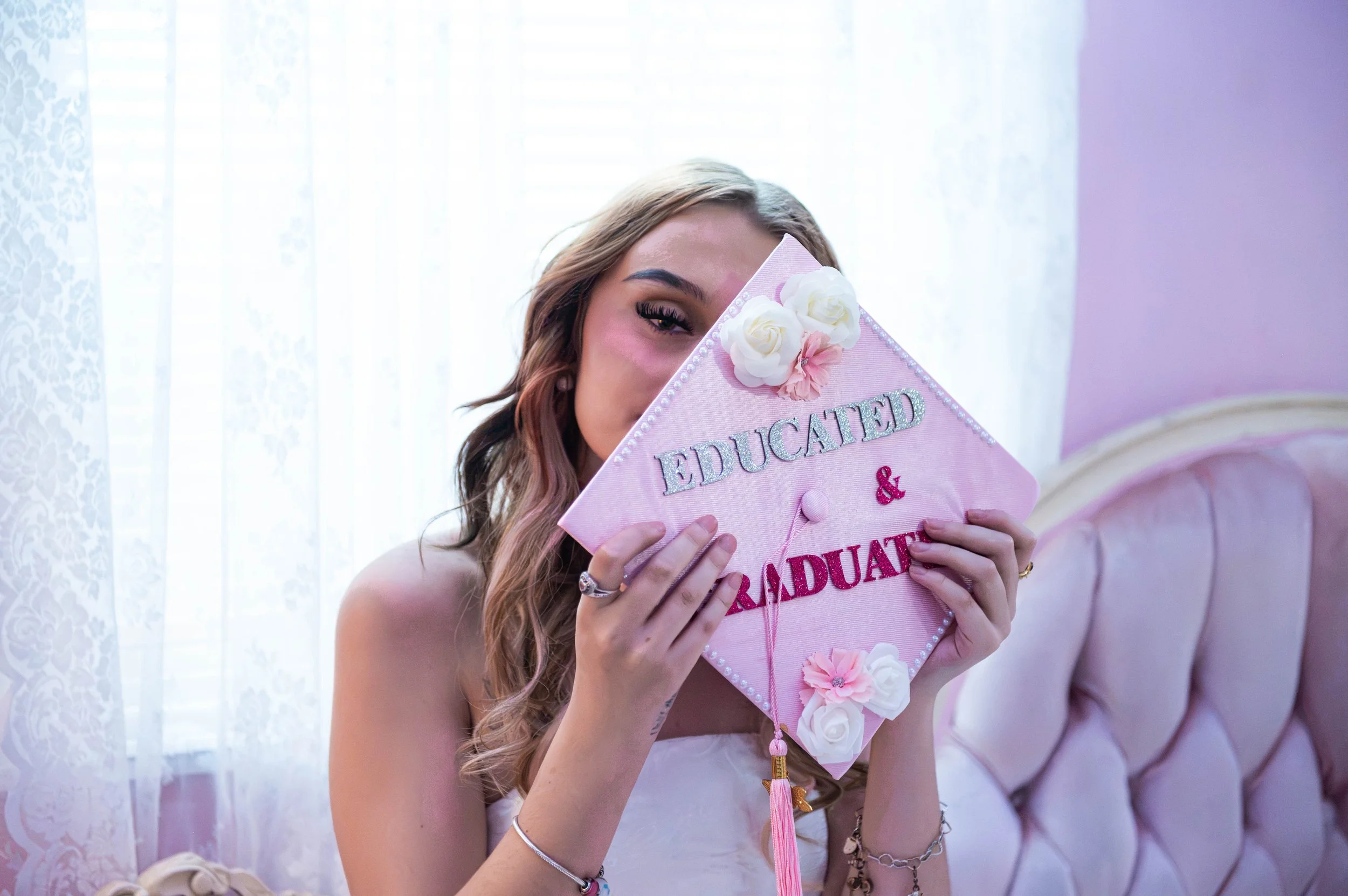 A woman with long wavy hair holding a pink graduation cap with white flowers and the words 'Graduated' on it, partially covering her face, smiling indoors with sheer white curtains and purple walls in the background.