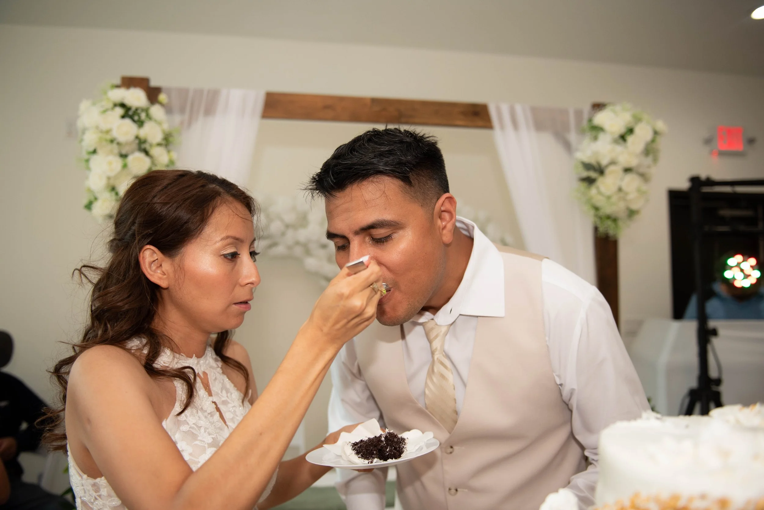 A woman is feeding a man a piece of cake at a wedding reception with floral decorations in the background.