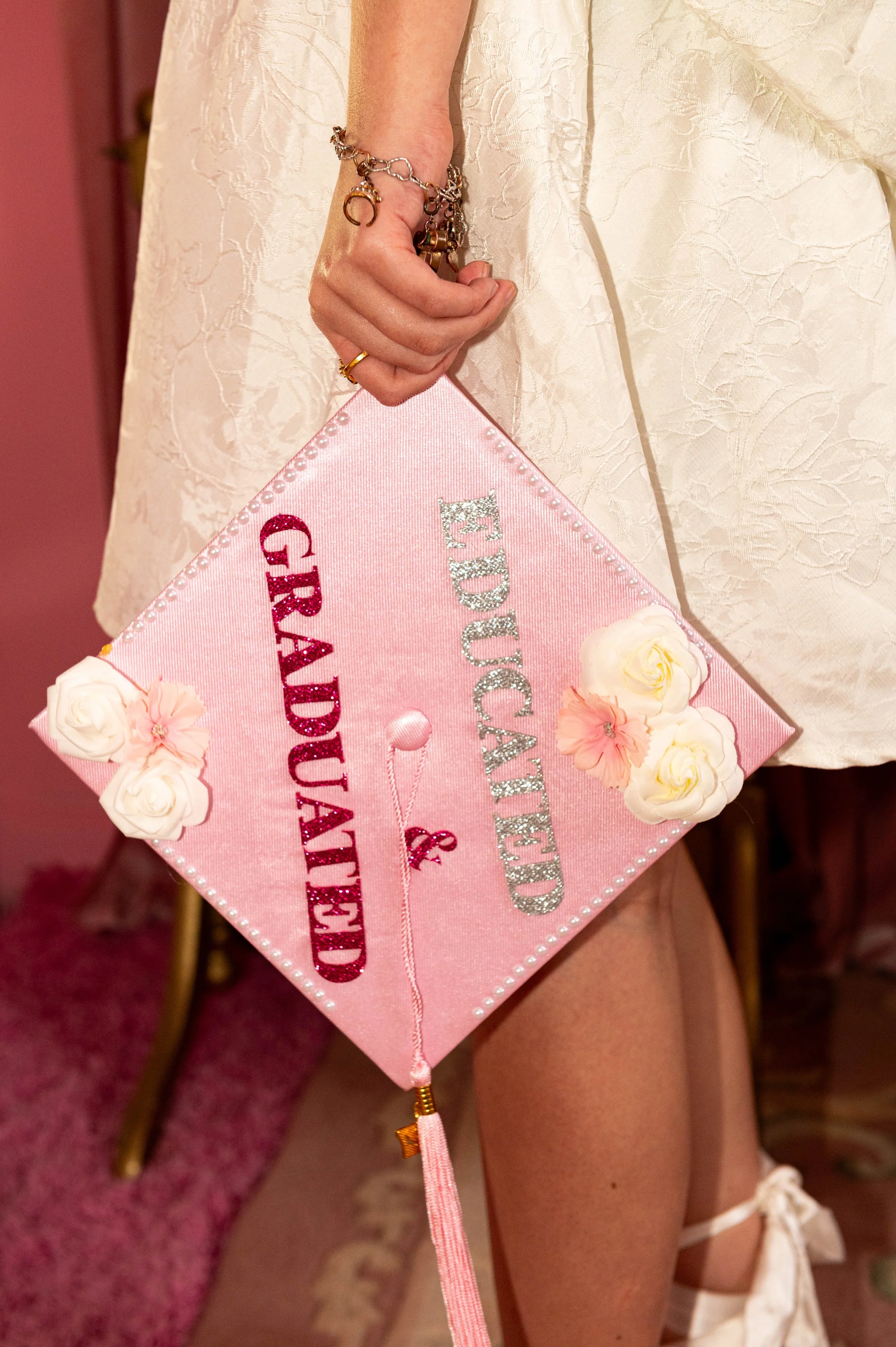 Close-up of a person's hand holding a pink satin pillow with embroidered text and flowers, wearing jewelry including a ring and bracelets, with a cream-colored textured dress in the background.