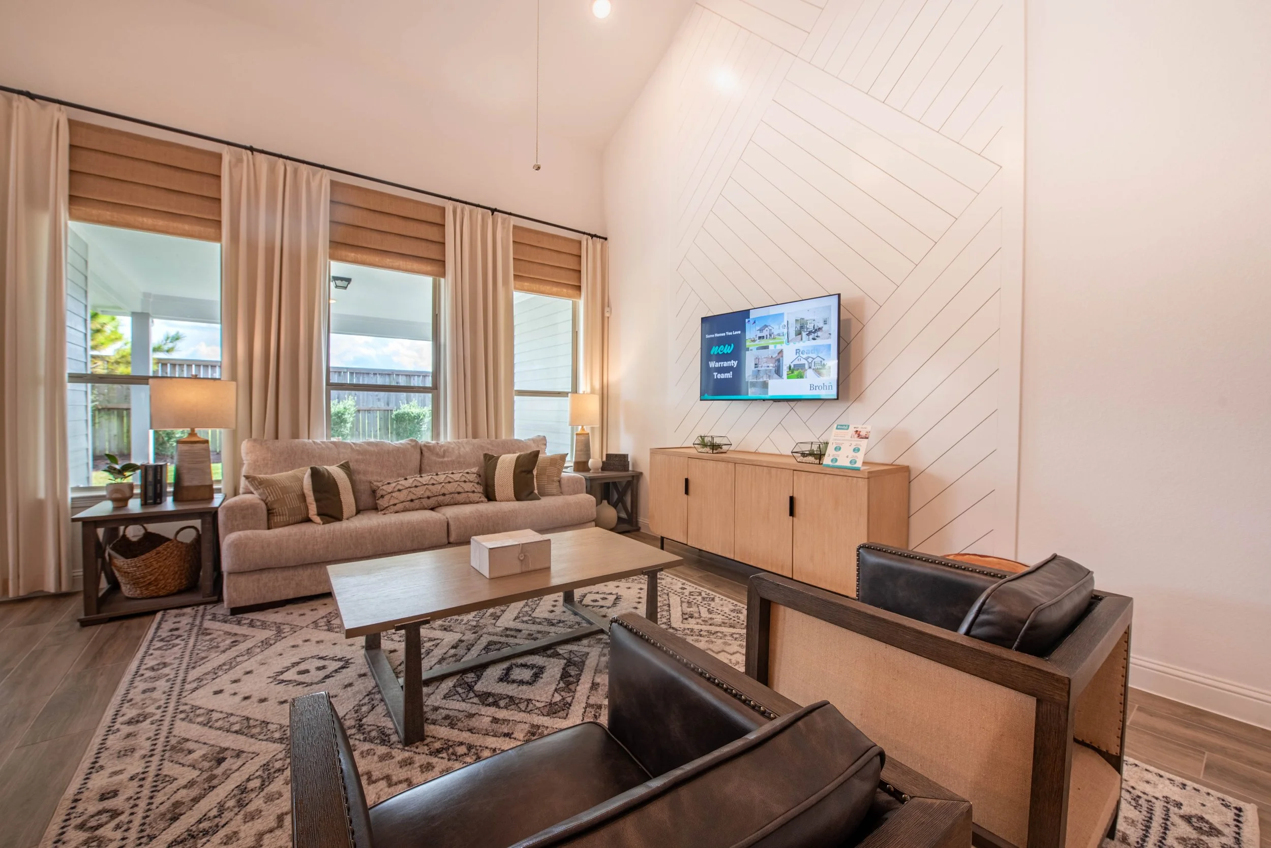 Living room with large window and beige curtains, a beige sofa with multiple cushions, a wooden coffee table, black and tan armchairs, a patterned rug, a wall-mounted TV, a wooden cabinet, and lamps on side tables. Outdoor patio and sky visible throu