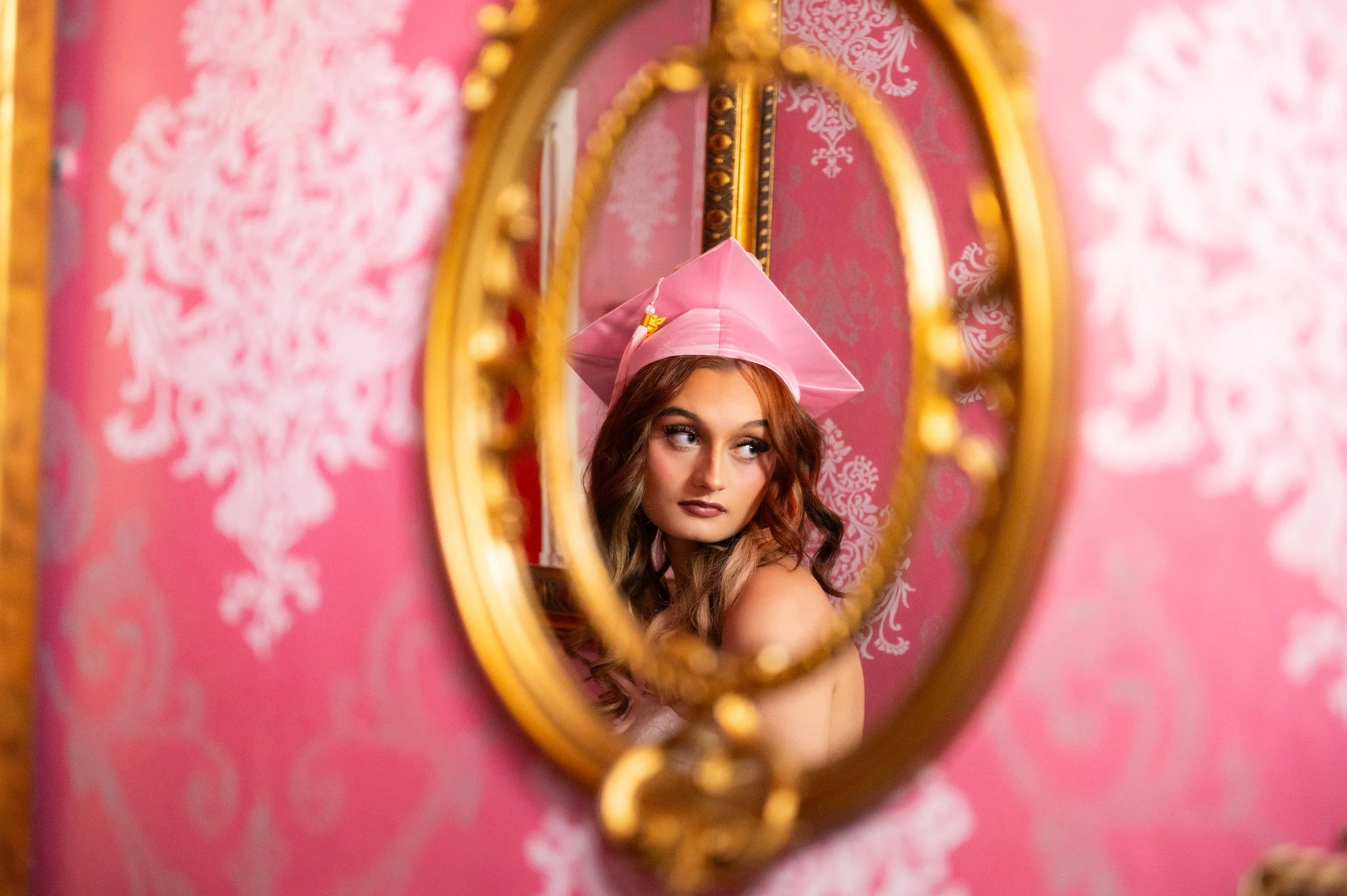 Woman looking into an ornate oval mirror with gold rim, pink hat, long wavy hair, pink wall with white floral pattern in the background.